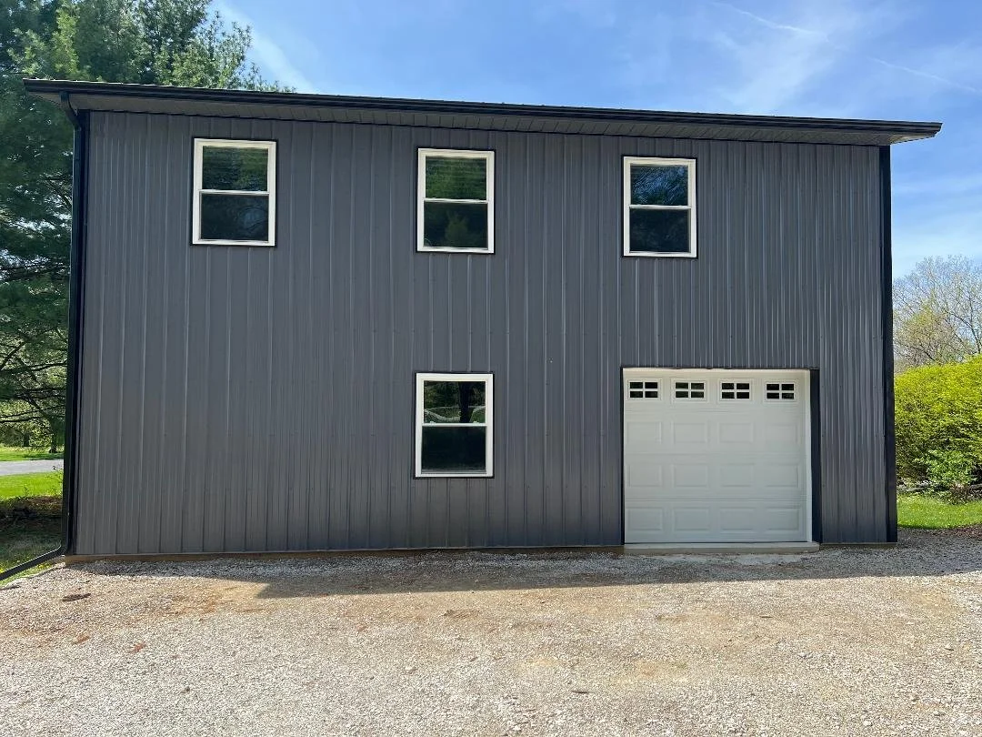 A two-story building with gray metal siding, four windows on the front with white trims, and a white garage door. The building is set on a gravel surface with trees and a blue sky in the background.