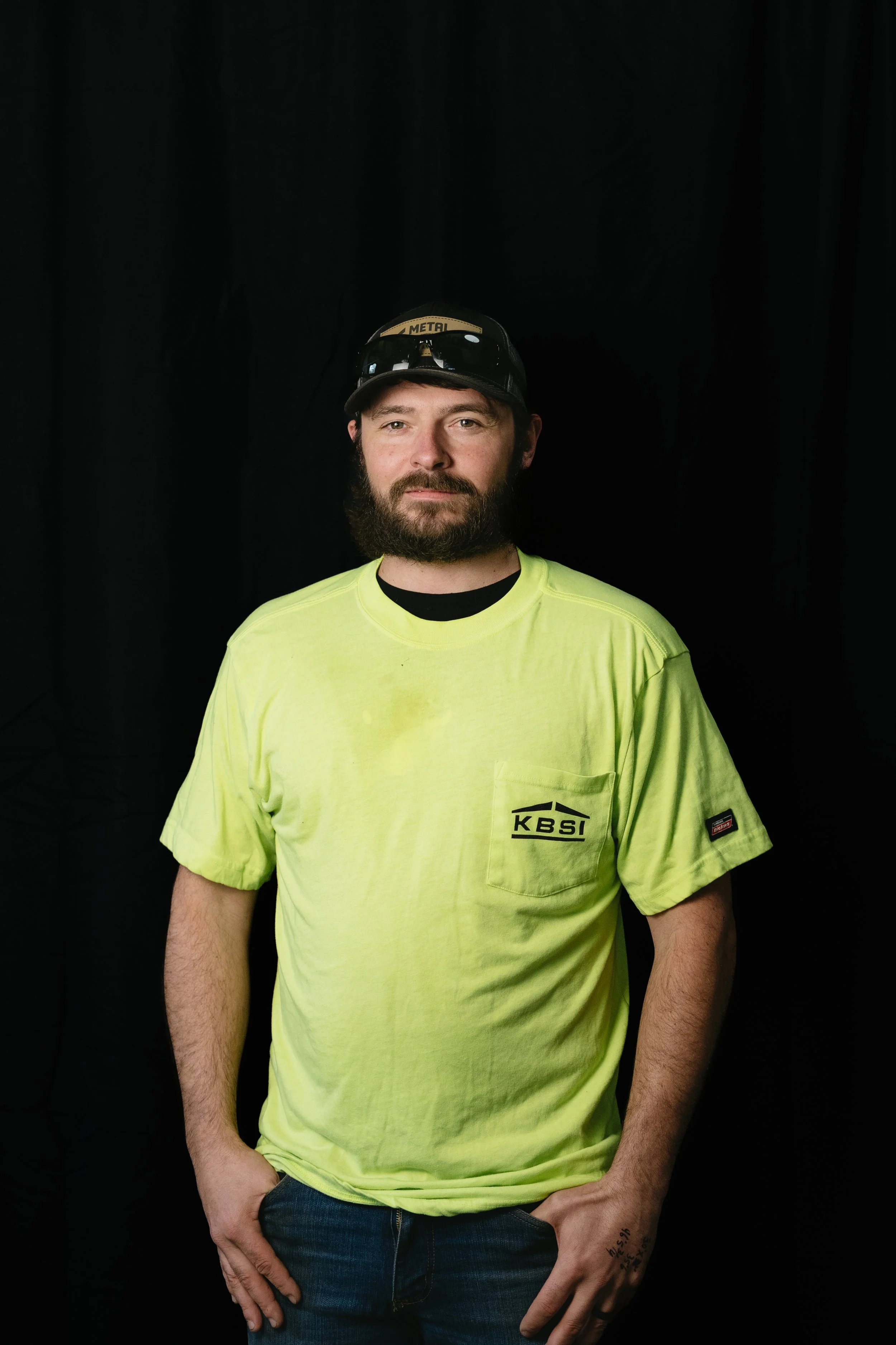Portrait of a man with a beard and mustache wearing a high-visibility yellow T-shirt with 'KBSI' logo, a baseball cap with 'METAL' label, and sunglasses on his head, standing in front of a black background.