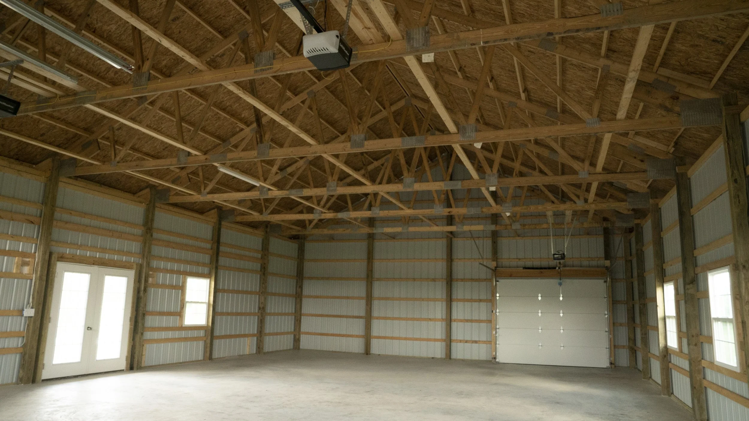Interior view of an unfinished garage framed with wood, showing exposed roof trusses and a concrete floor, with a garage door at the back and windows along the side walls.
