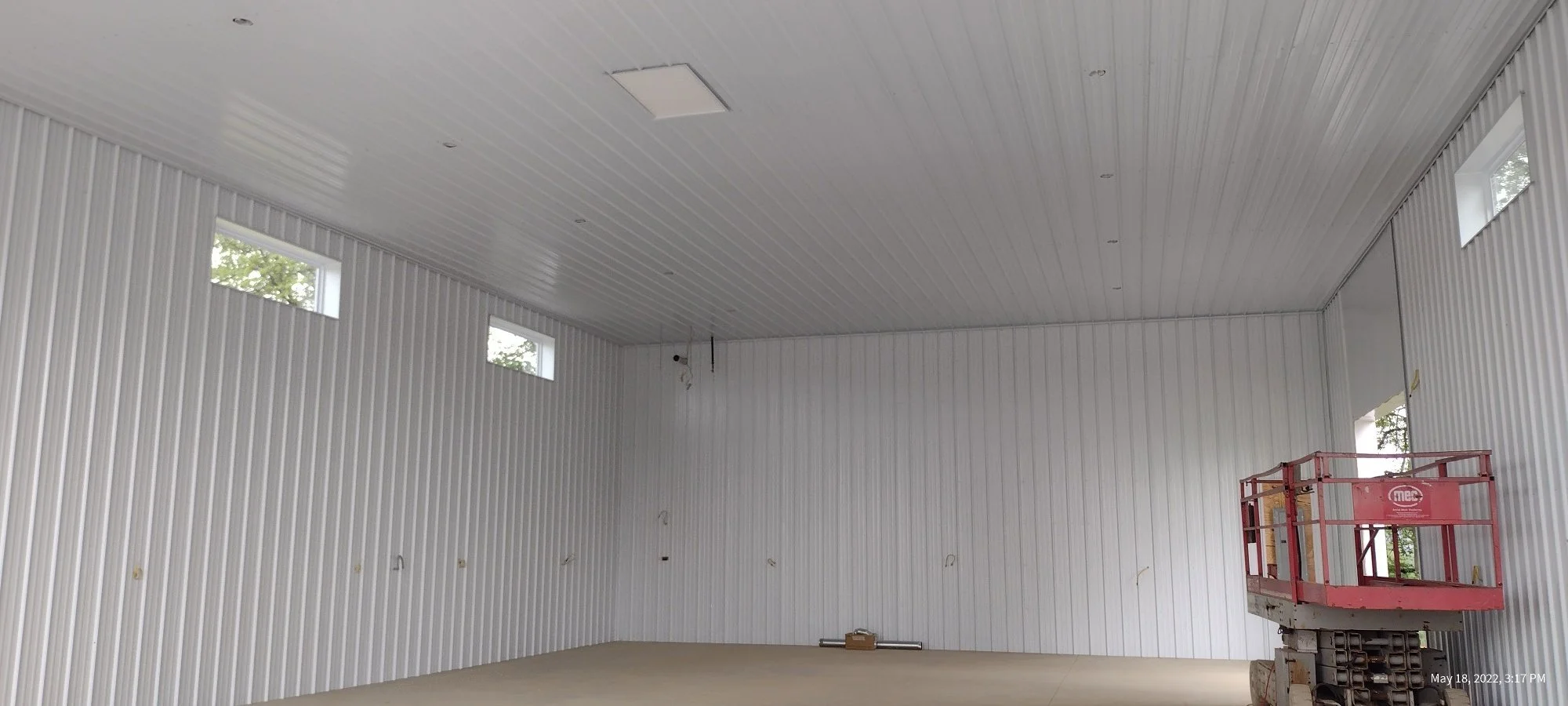 Interior of a large, white steel-paneled building under construction, with high walls and ceiling, small rectangular windows, and a red scissor lift in the corner.