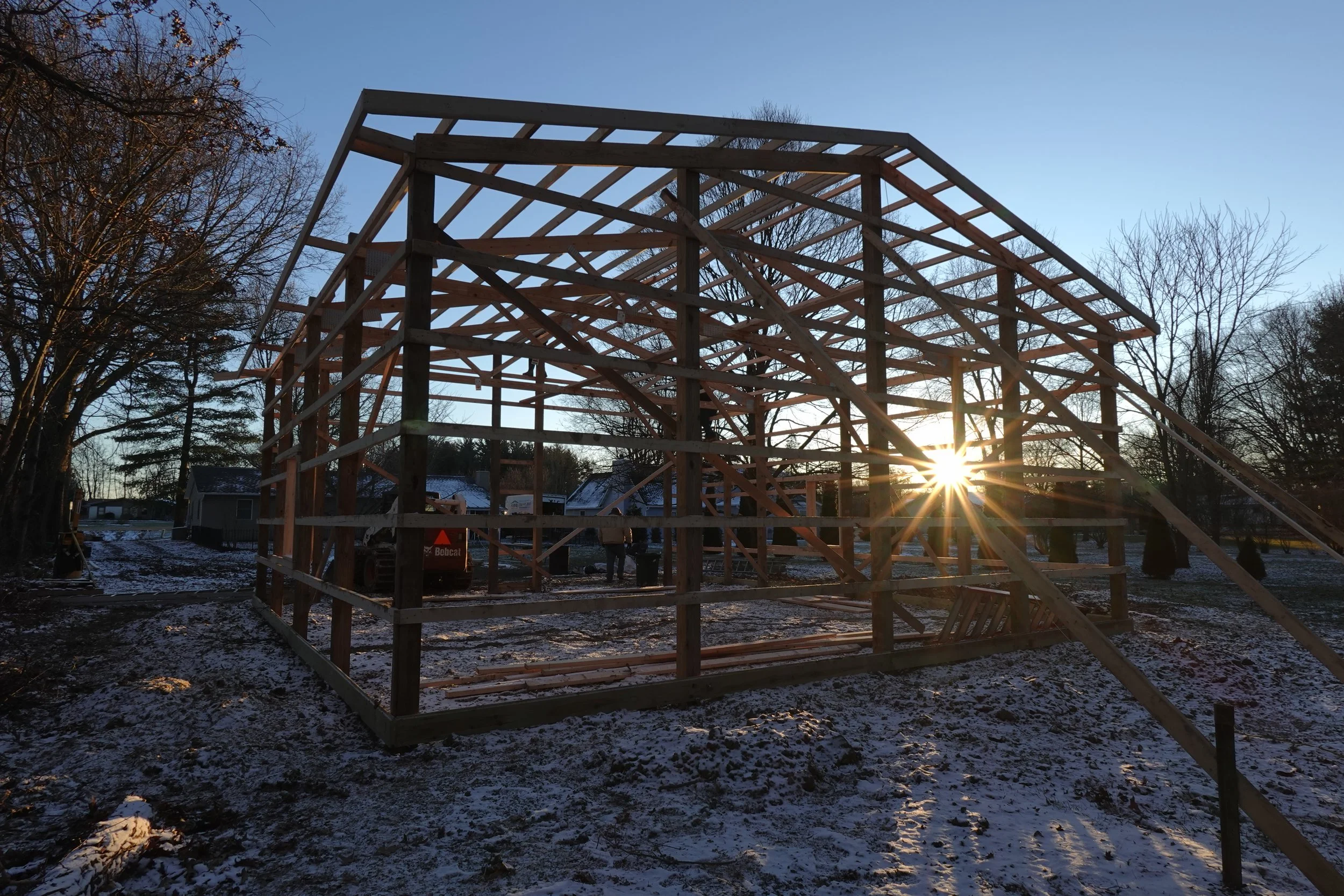 Wooden framing of a house under construction at sunset, with trees and snow on the ground.