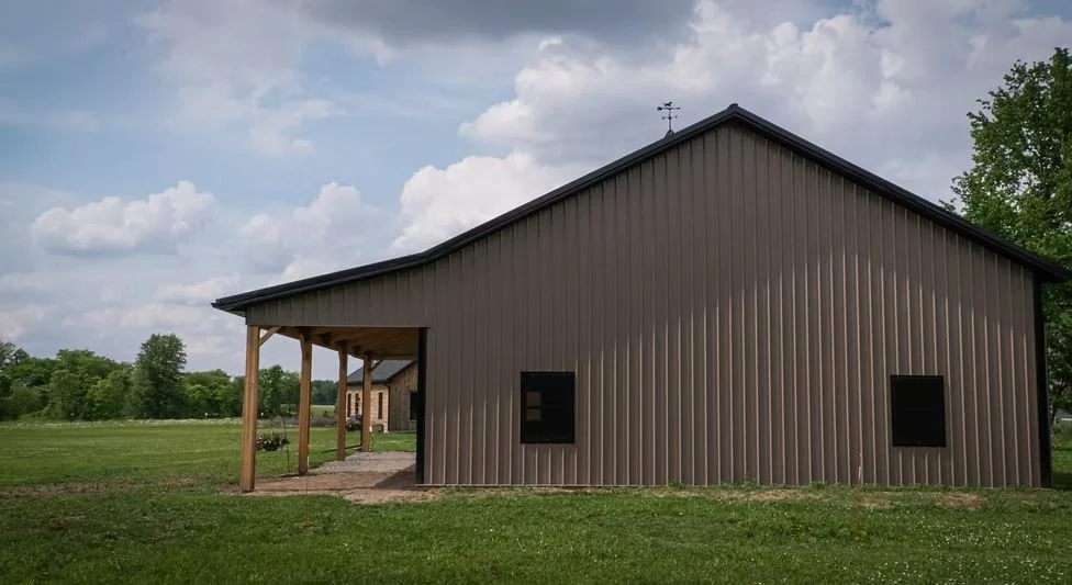 A metal building with a sloped roof, two small black windows, and wooden support beams on a grassy landscape under a partly cloudy sky.