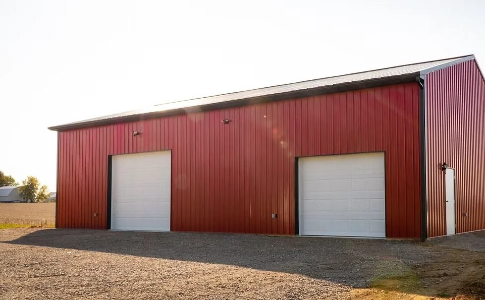 Red metal warehouse building with two large white garage doors and a side door, situated on a gravel lot with open land in the background.