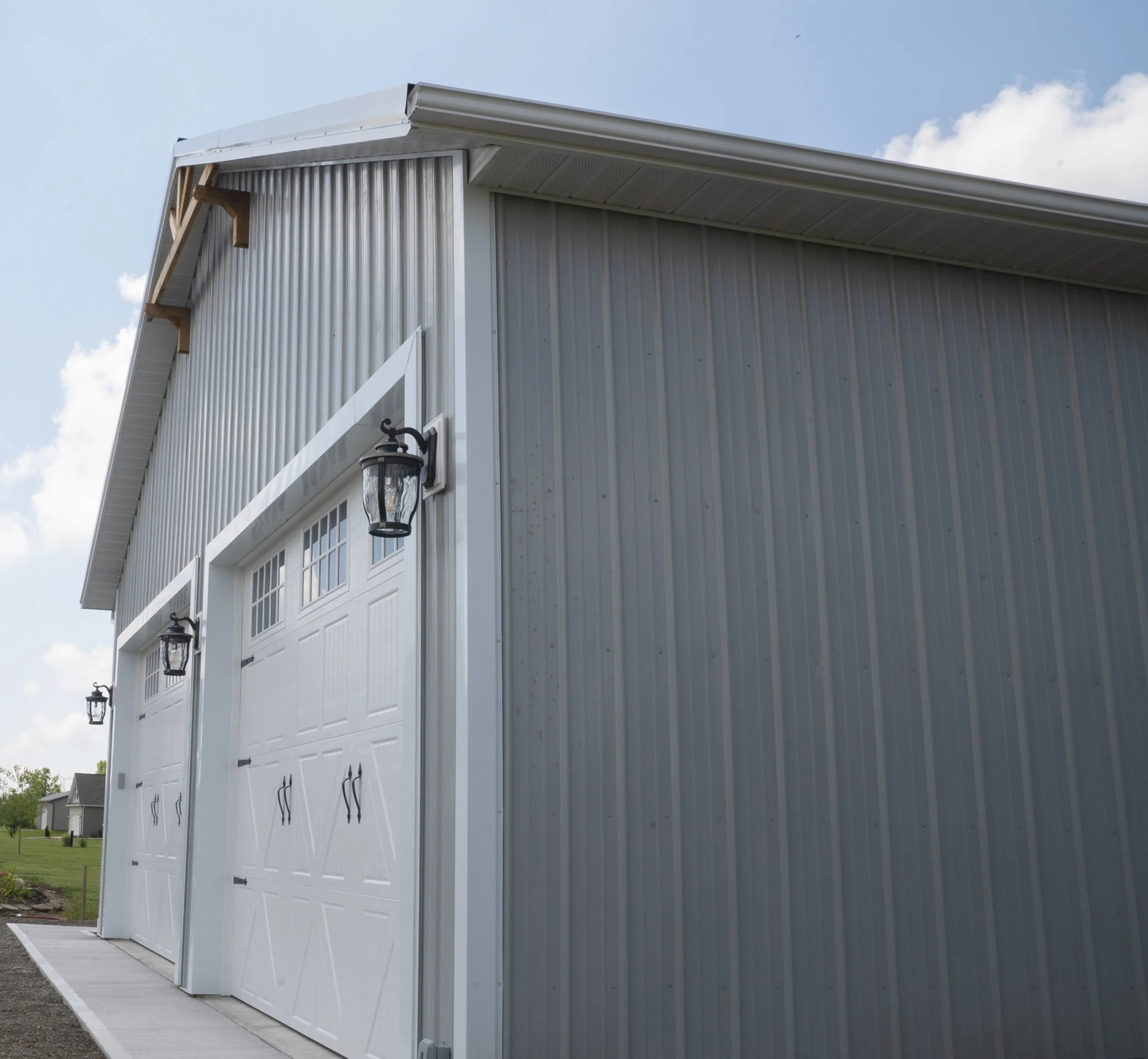 Large silver metal building with white garage doors, black lantern-style lights, and a sloped roof with wooden supports visible at the corner against a blue sky with a few clouds.