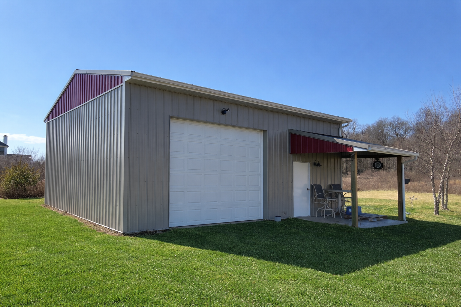 Metal storage shed with a large garage door, a side door, and a covered porch area with chairs and a small table, set on a grassy lawn under a clear blue sky.