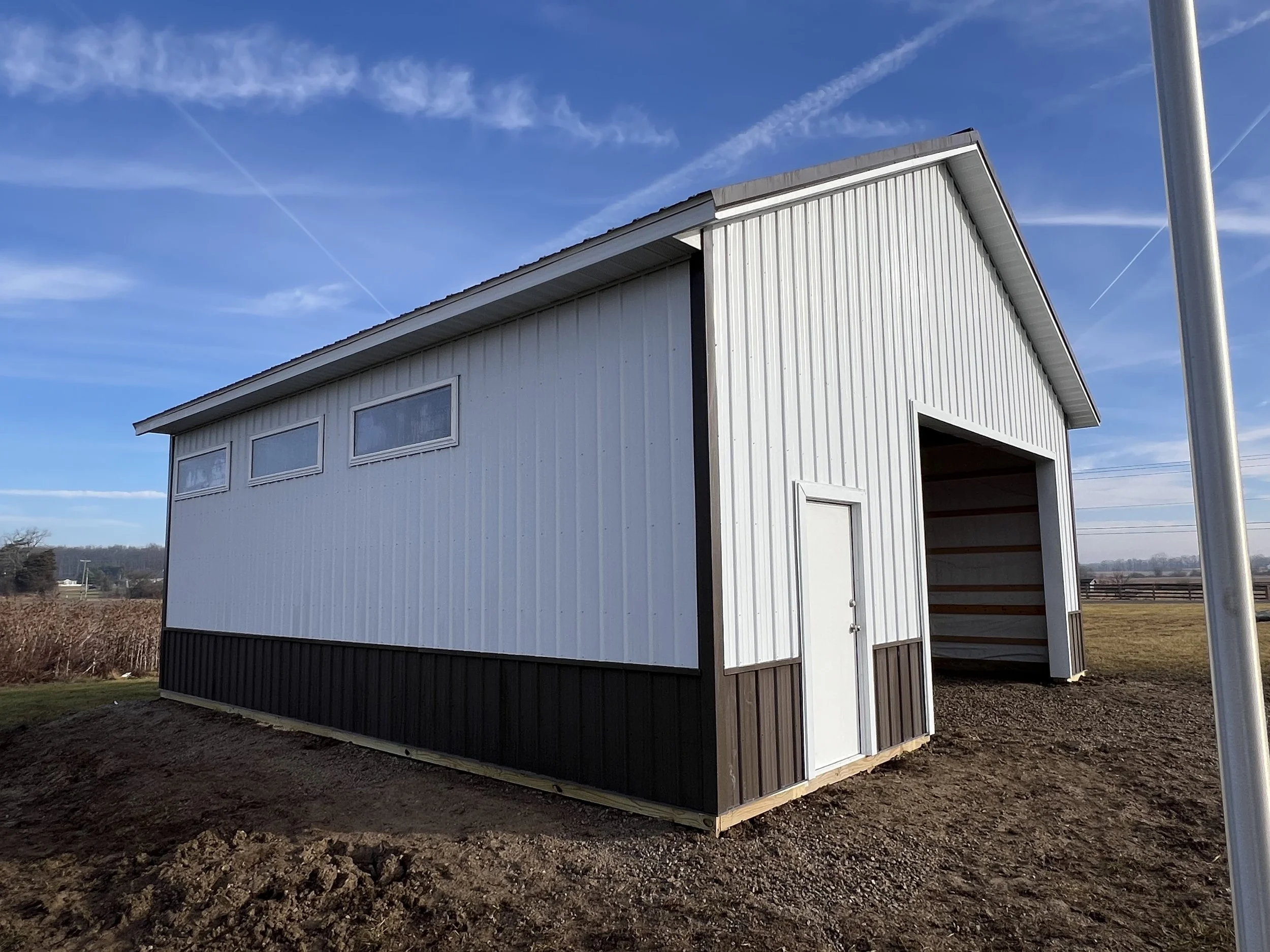 A white metal building with black trim under a blue sky with scattered clouds. The building has multiple windows and a large open garage door. The ground around the building appears to be dirt and gravel. In the background, there are fields and some 