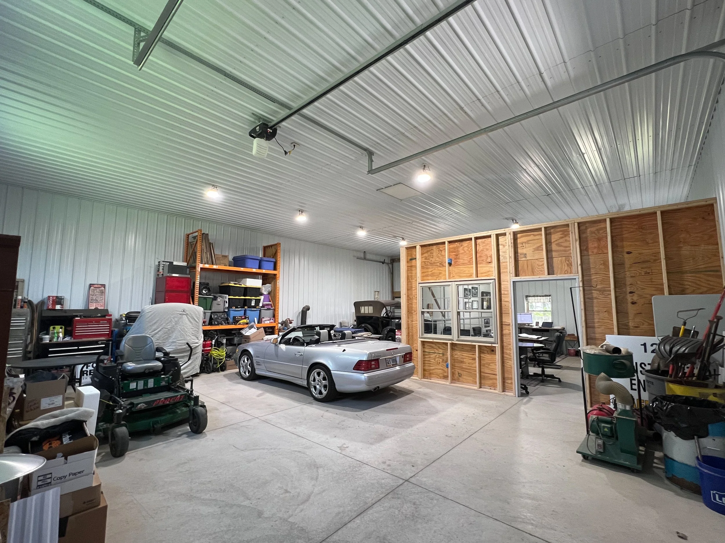 Interior of a large garage or workshop with a metal ceiling, fluorescent lights, various tools and storage shelves, a silver car in the center, and unfinished wooden wall framing on the right side.