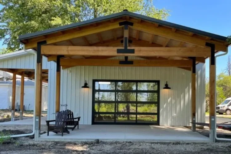 Front view of a modern garage or workshop with a large glass door, wooden porch roof, two black wall-mounted lights, and two black chairs outside.