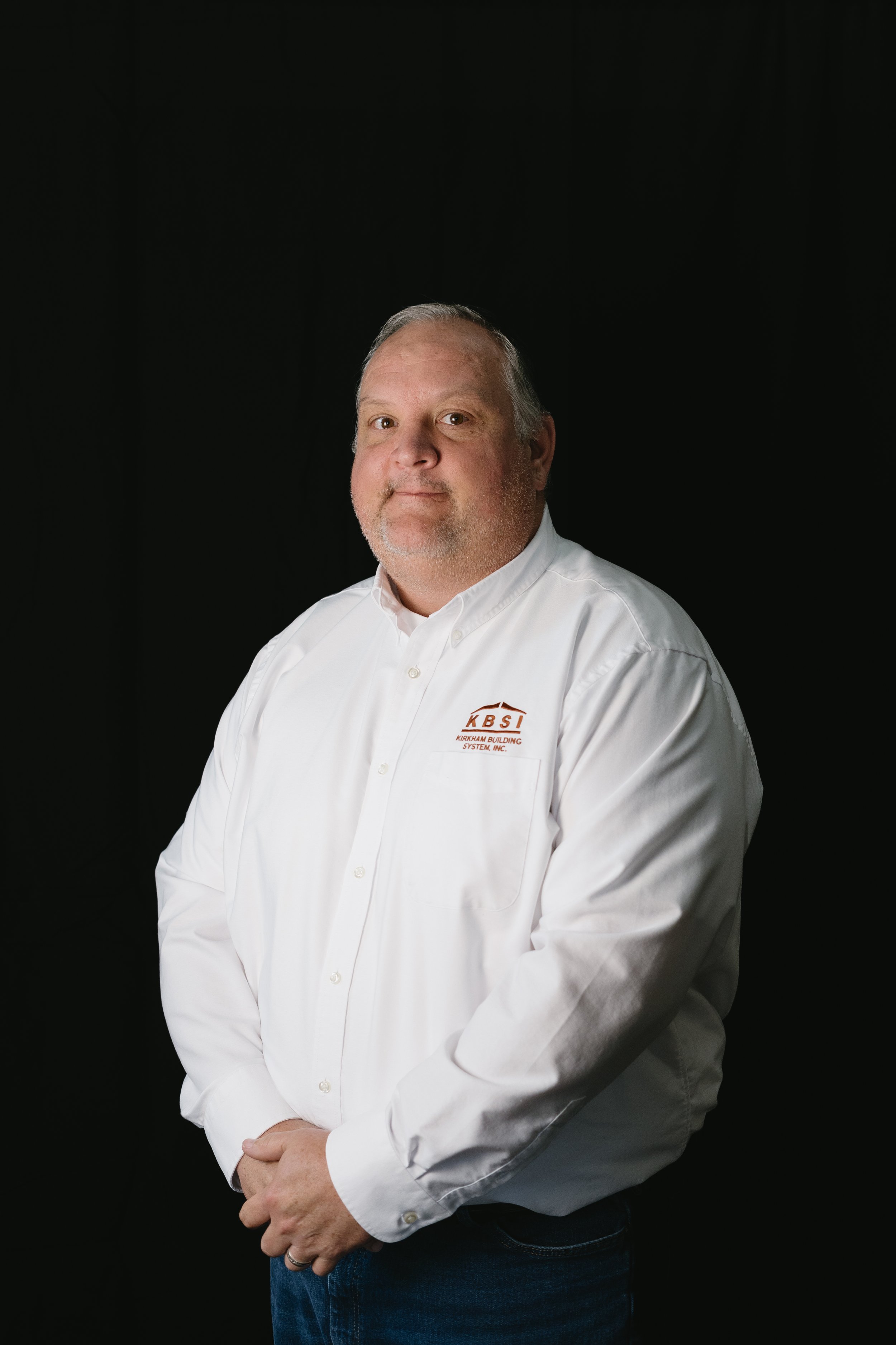 Portrait of a middle-aged man with short gray hair, wearing a white button-down shirt with a logo that reads 'KBSI' and dark jeans, standing against a black background.