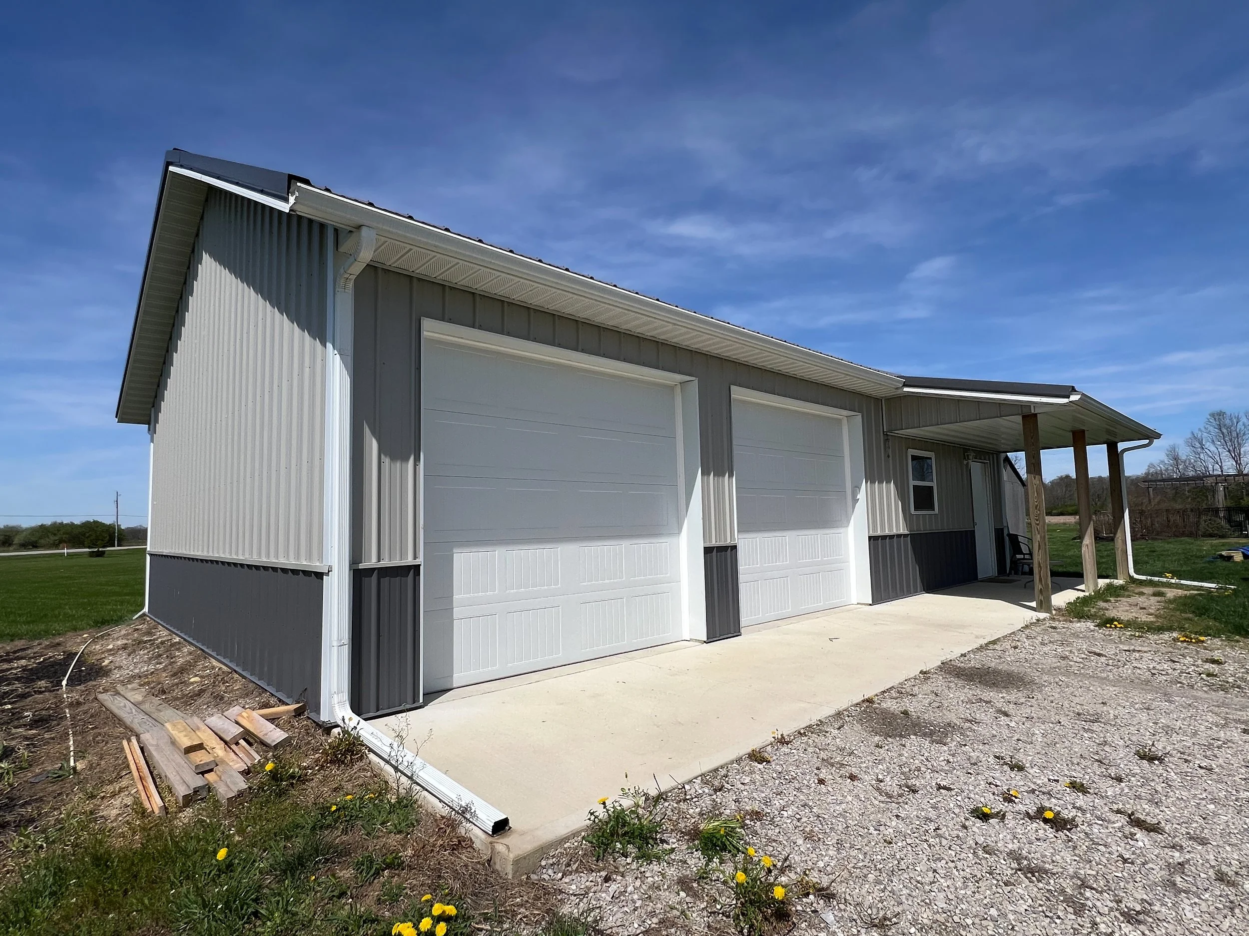 A white garage with closed white doors, metal siding, and a concrete driveway outdoors on a sunny day with a blue sky and some trees in the background.