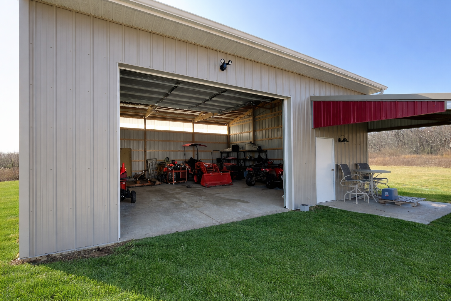 A metal garage with an open door revealing various outdoor equipment and a lawn outside. Next to the garage, there is an outdoor seating area with chairs, a table, and some items on the ground.