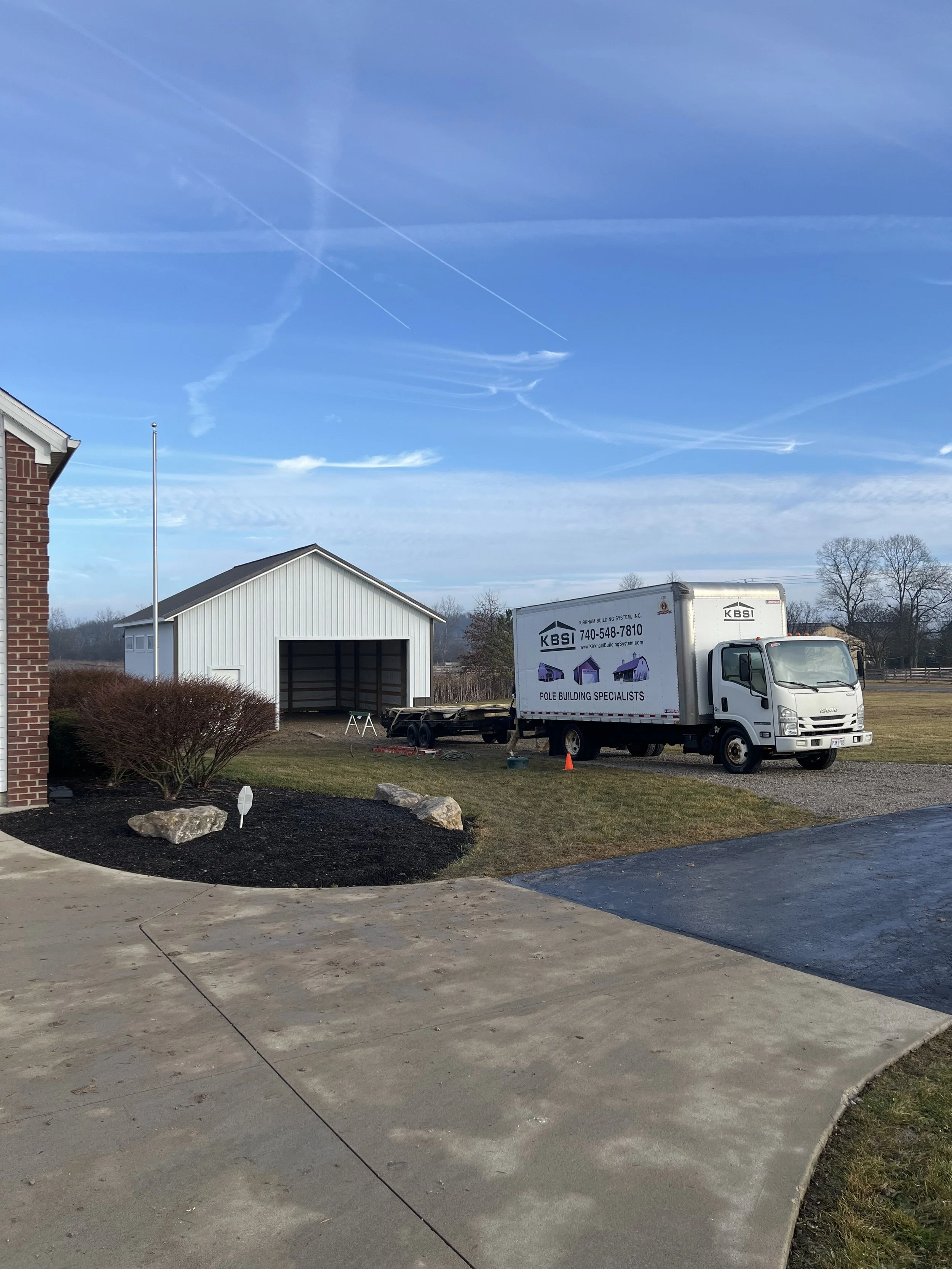 A suburban scene with a white garage, a moving truck labeled 'KBSI Pole Building Specialists,' a brick house with an American flagpole, and leafless trees in the background, under a partly cloudy sky with contrails.