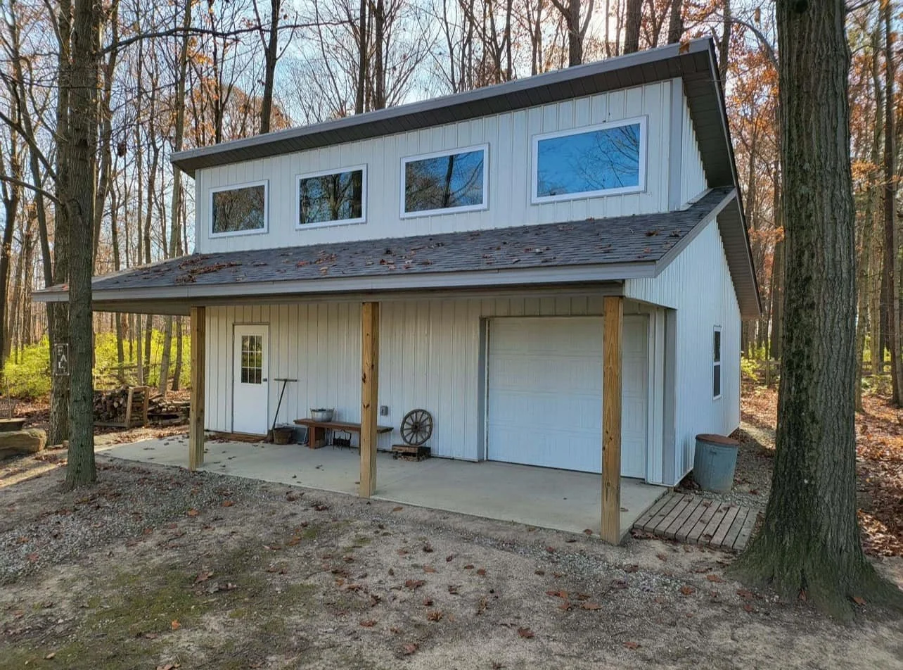 A two-story white barn-style building with a garage door and a small side door, situated in a wooded area during fall.