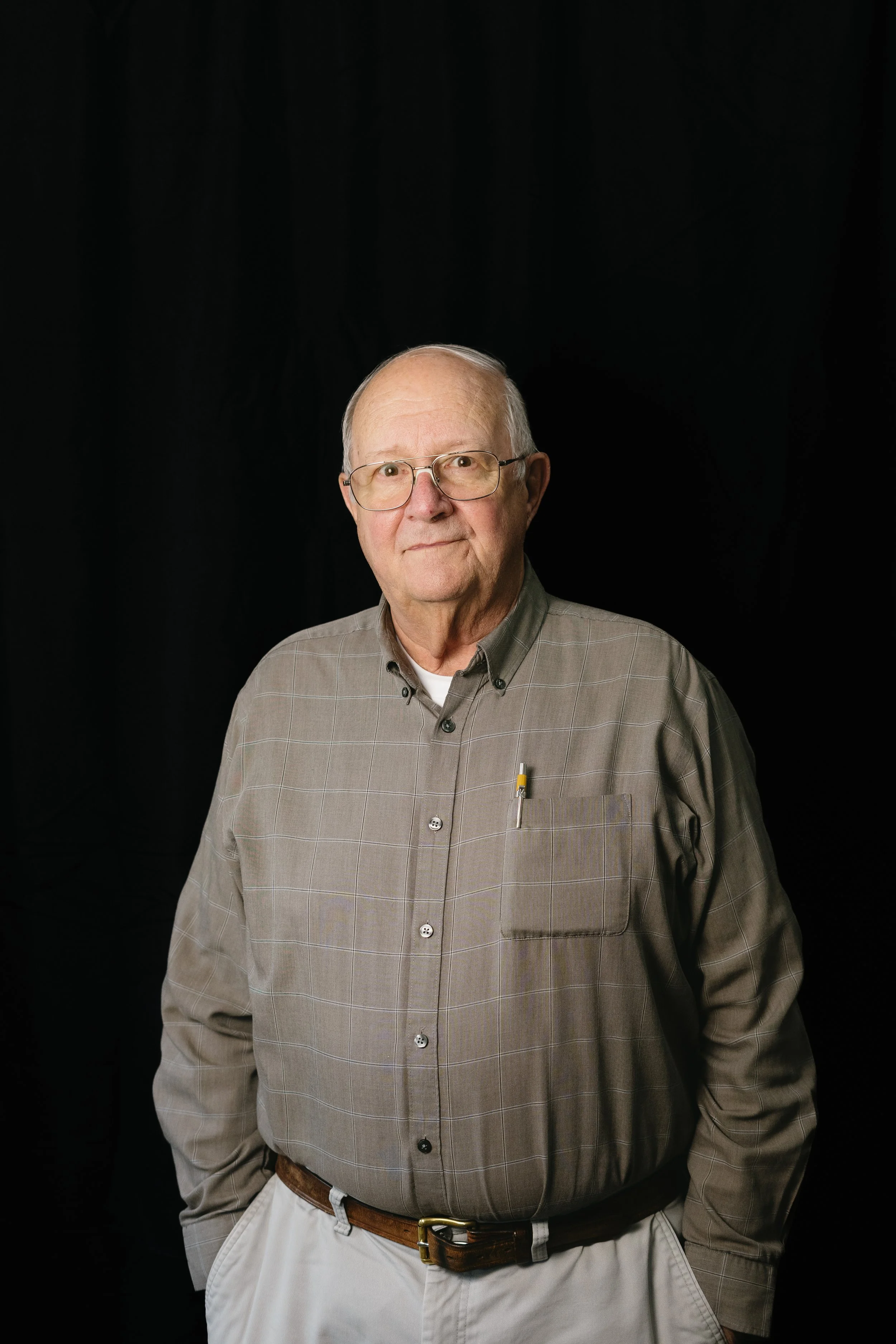 A portrait of an elderly man with glasses, wearing a plaid shirt, standing against a black background.