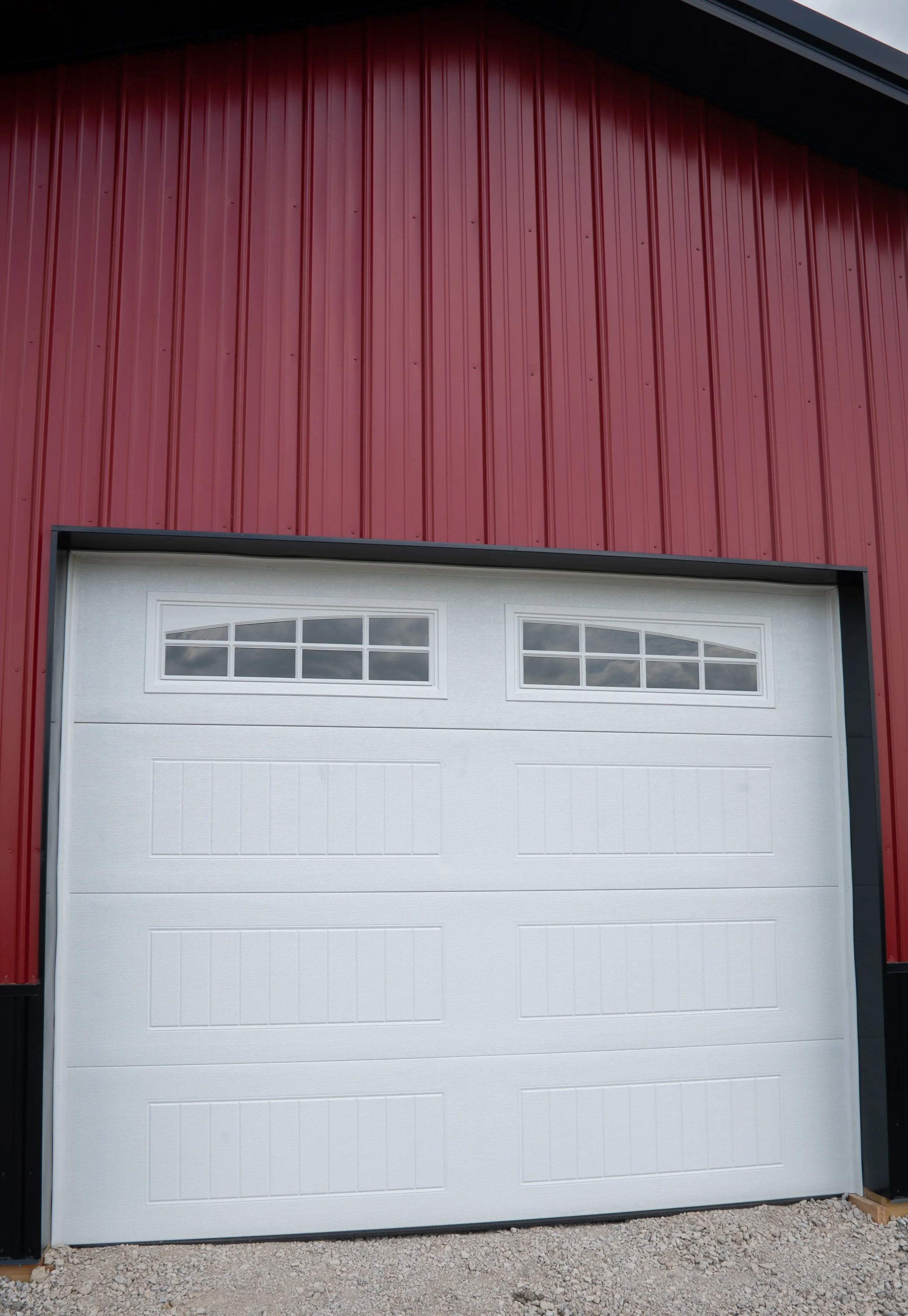 A white garage door with rectangular windows near the top, installed in a red metal building with vertical paneling, on a gravel ground.