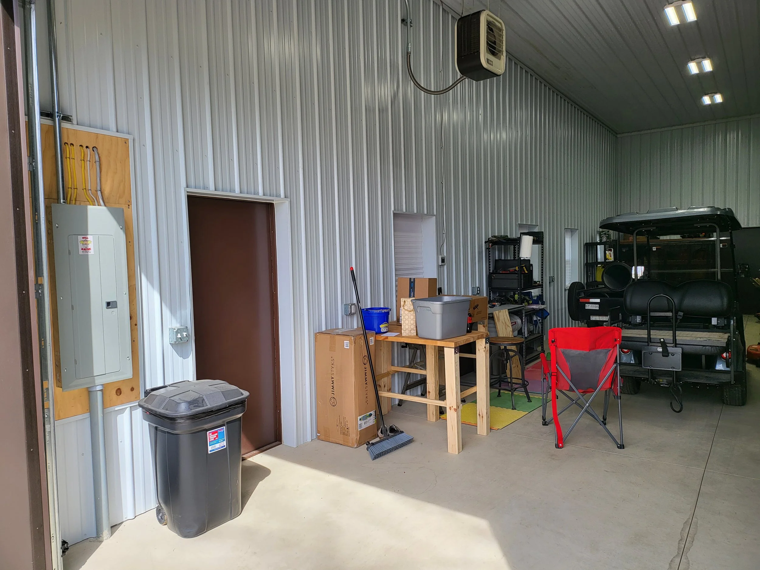 Inside a garage with metal siding, various household and yard items including a trash bin, cardboard boxes, a table, shelving units, and outdoor equipment such as a golf cart or small utility vehicle.