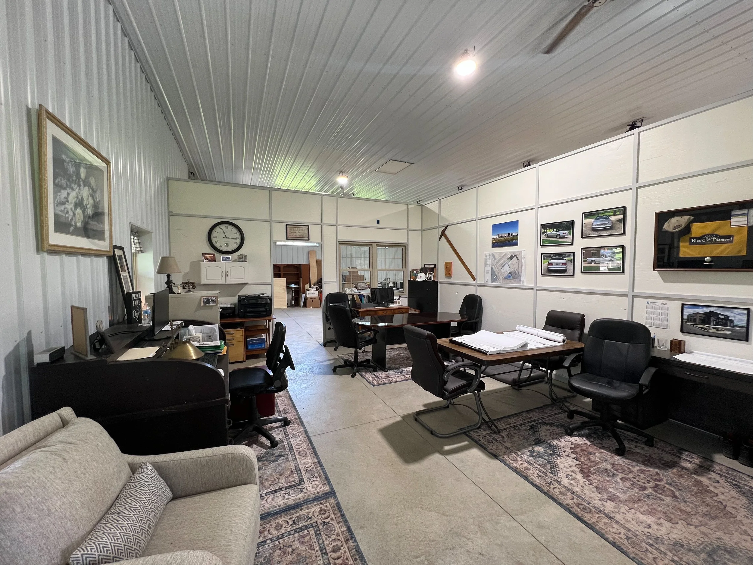 An office with multiple desks and chairs, framed pictures of cars on the wall, and a clock. There's a beige couch with a patterned pillow in the foreground, and various papers and office supplies on the desks.