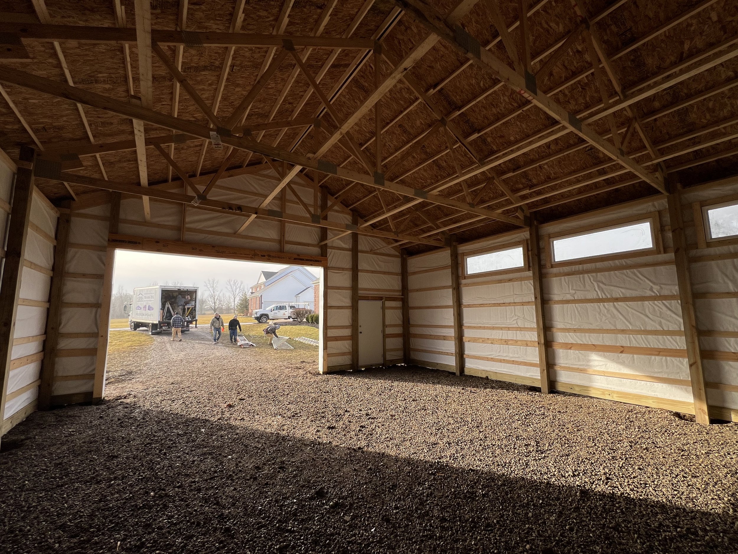 The interior of a building under construction with exposed wooden framing and trusses, large open garage door, and several construction workers outside.