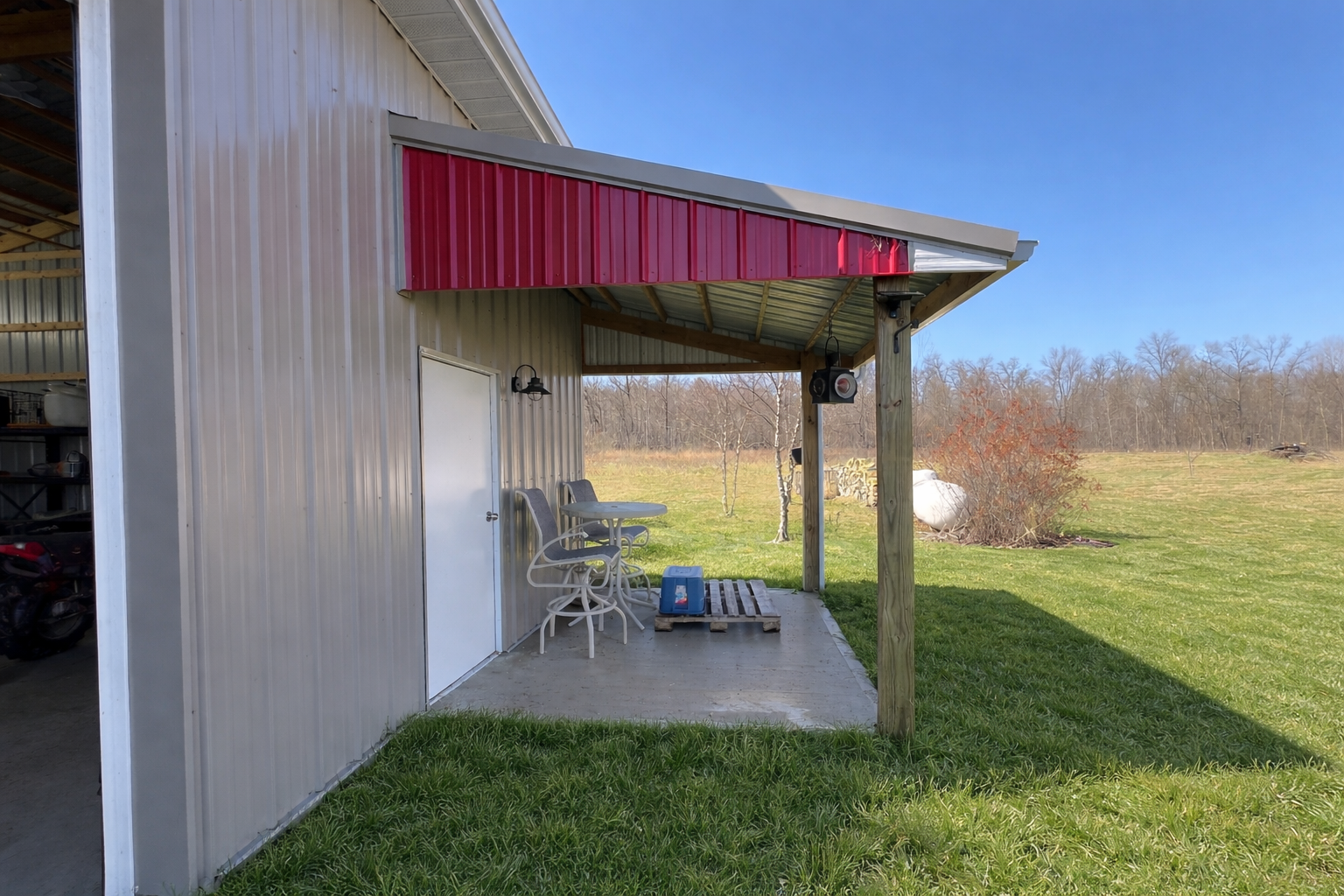 Backyard shed with metal walls featuring a red awning, white door, two chairs, a small table, and a blue cooler, with a grassy yard and trees in the background under a blue sky.