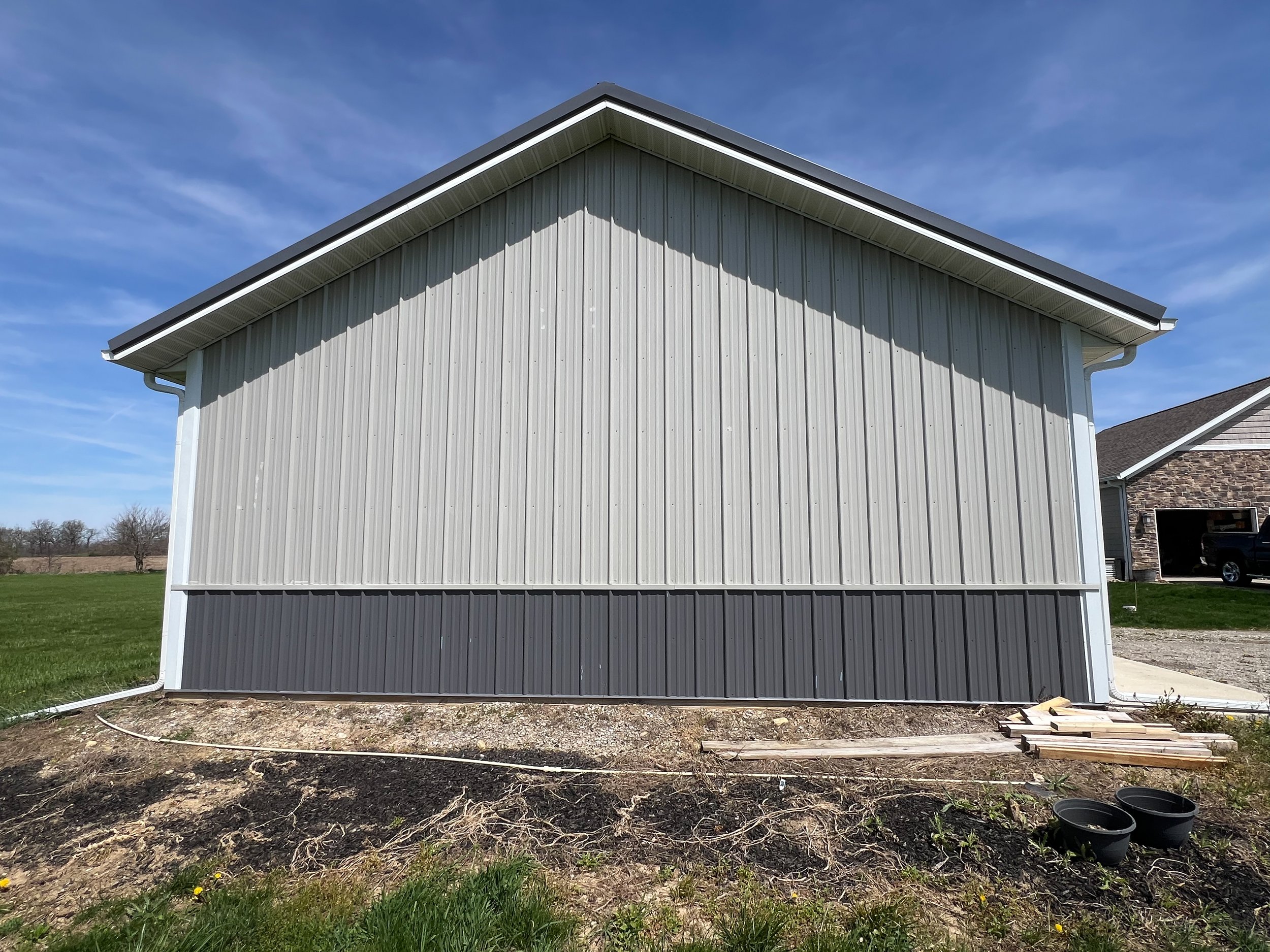 Side view of a white sliphouse with vertical siding, under construction with an exposed ground area and construction materials nearby. The house has a gabled roof and a section of brick siding on one side.