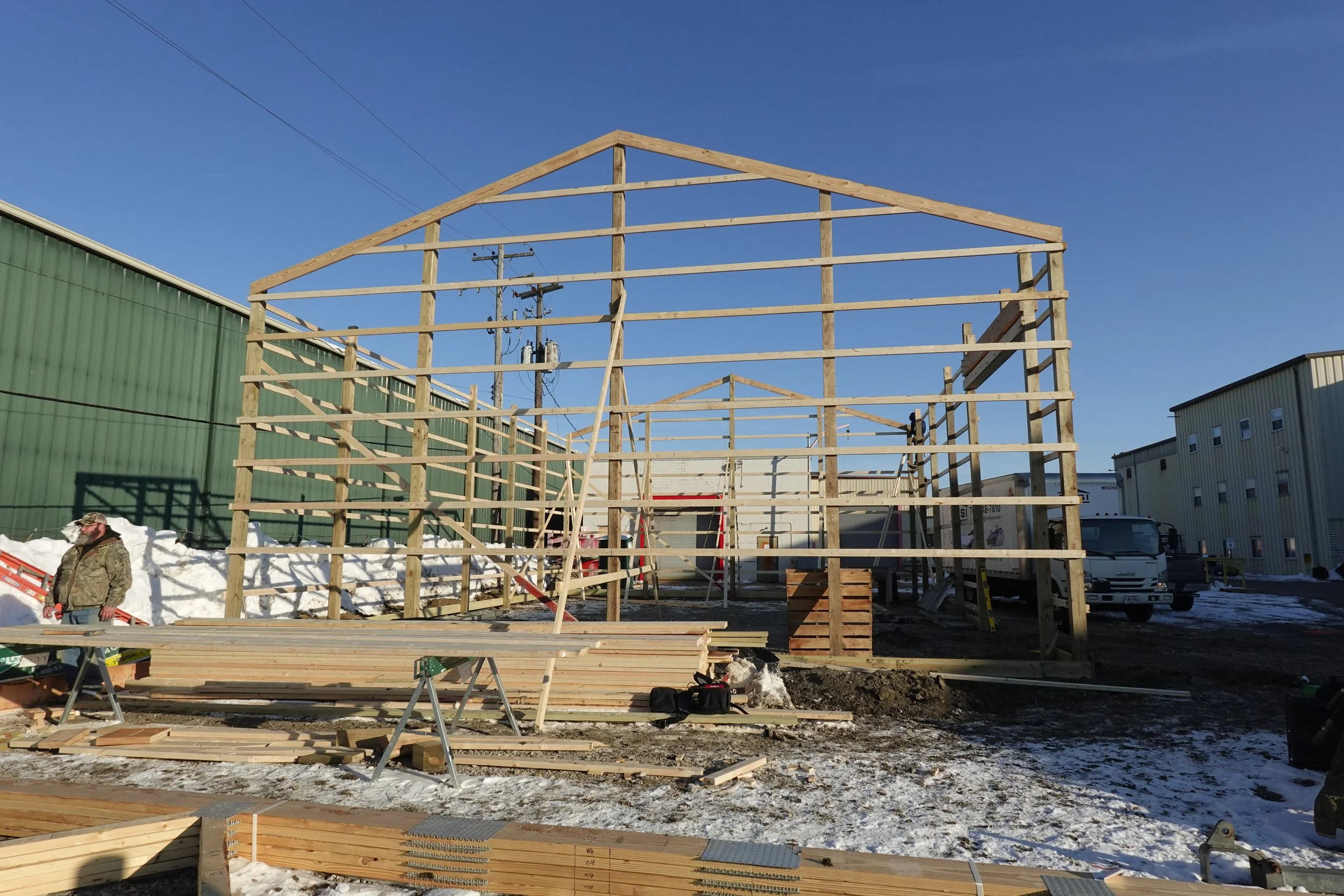 Construction site with a wooden frame structure being built, surrounded by industrial buildings and vehicles, with snow on the ground and a clear blue sky.