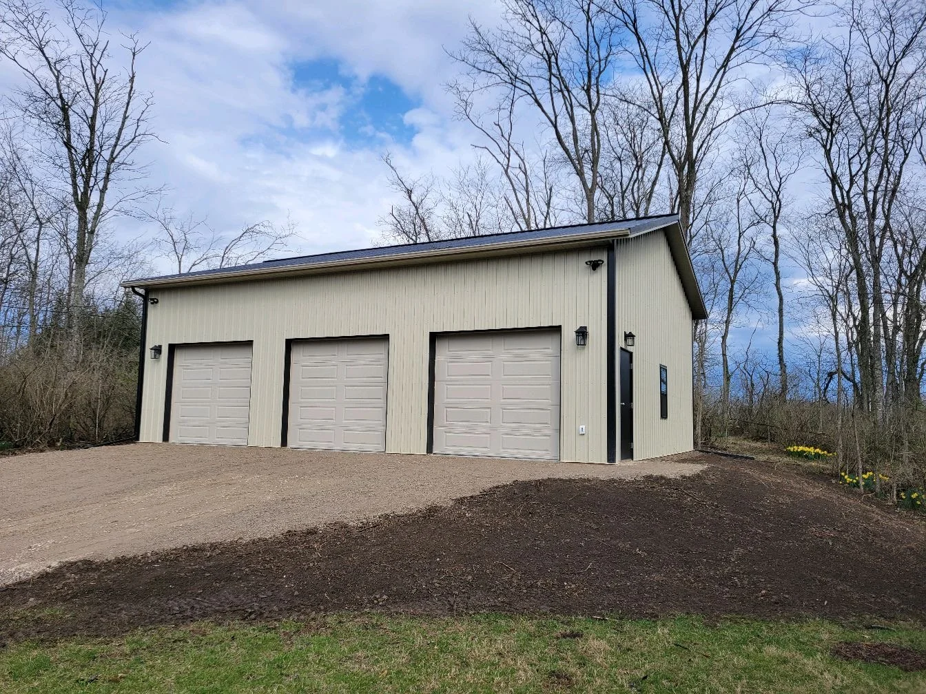 A large, modern garage with three beige garage doors, black trim, exterior lights, and a side entry door, surrounded by leafless trees and a dirt driveway.