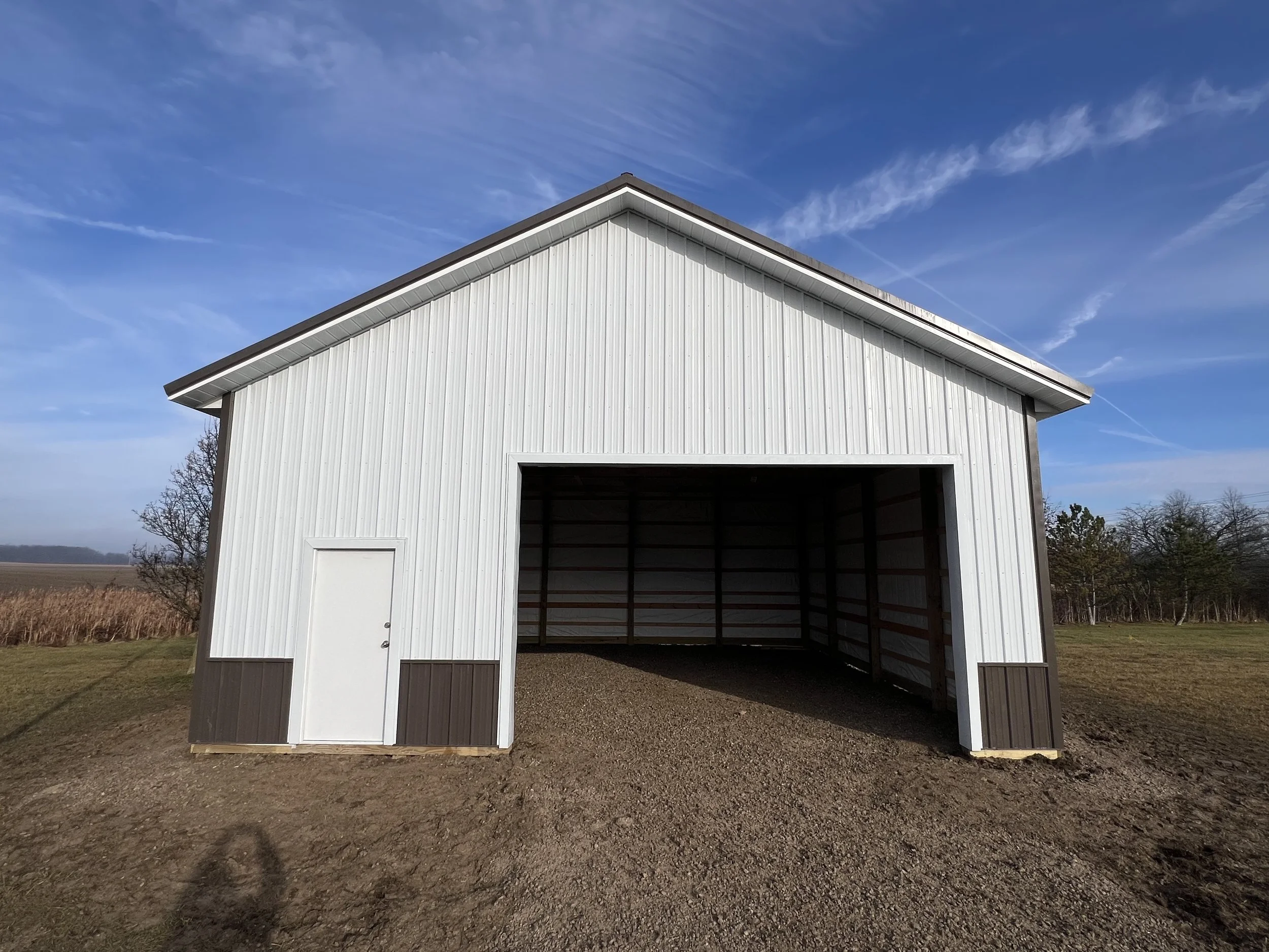 A white metal barn with an open front, standing on a gravel and grass field under a partly cloudy blue sky.