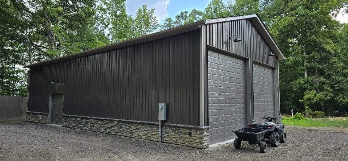A large, dark gray metal building with a stone foundation, two garage doors, and outdoor lighting, surrounded by trees and gravel ground.