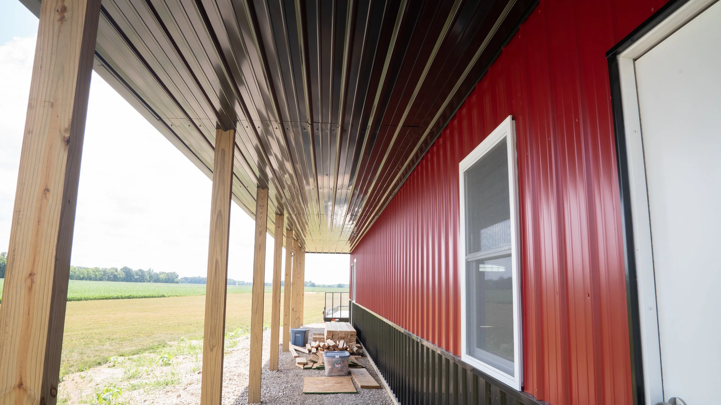 Under construction porch or deck attached to a red metal building with vertical siding, supported by wooden posts, with construction materials and tools on the ground.