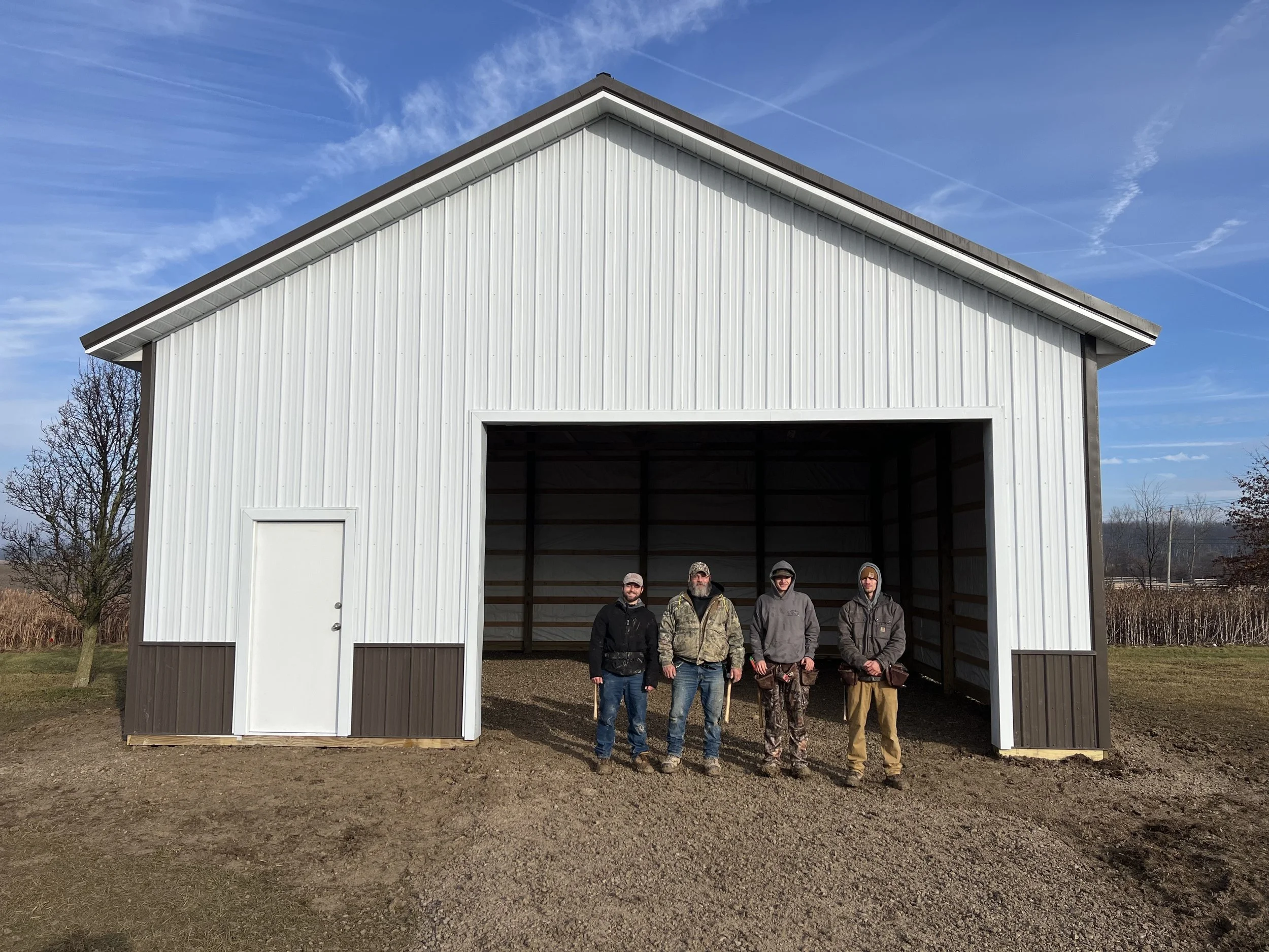 A group of four men standing in front of a large white metal building with a gabled roof, open garage door, and a smaller door on the side, on a cloudy day with a blue sky and trees in the background.