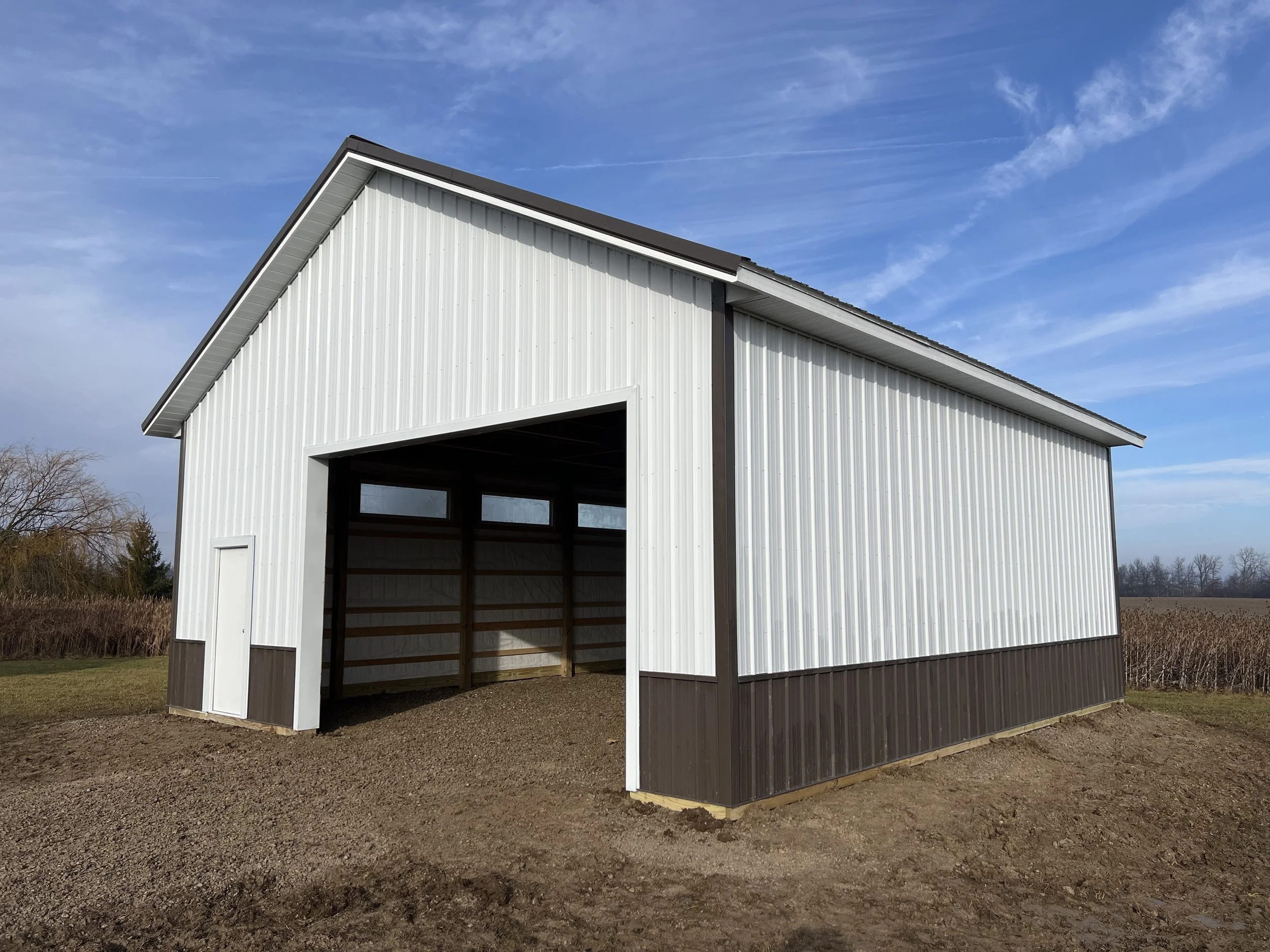 A large metal shed with an open door, situated on a dirt ground under a partly cloudy blue sky.