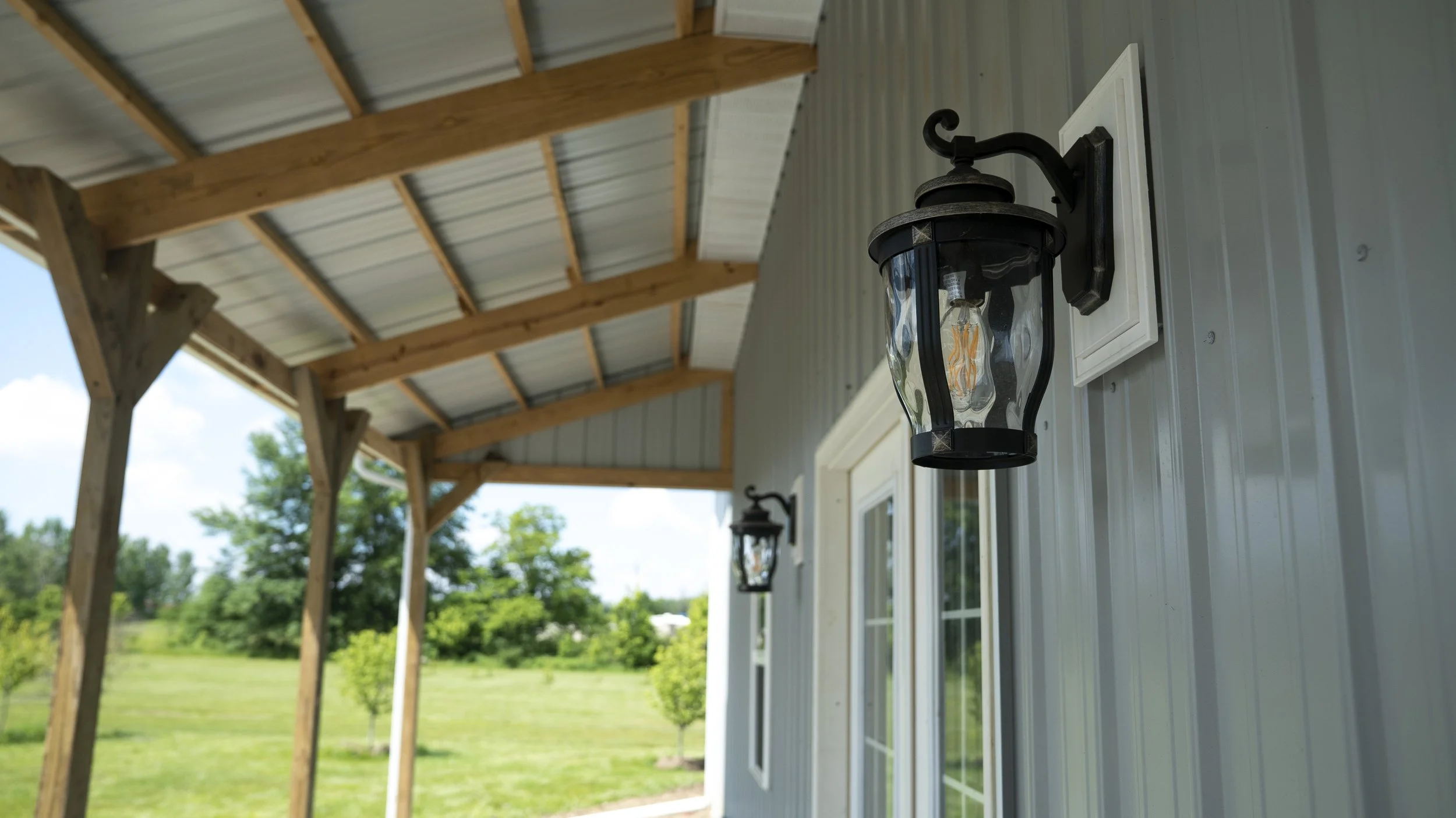 Outdoor porch with metal roof, wall-mounted lanterns, and view of green landscape with trees.