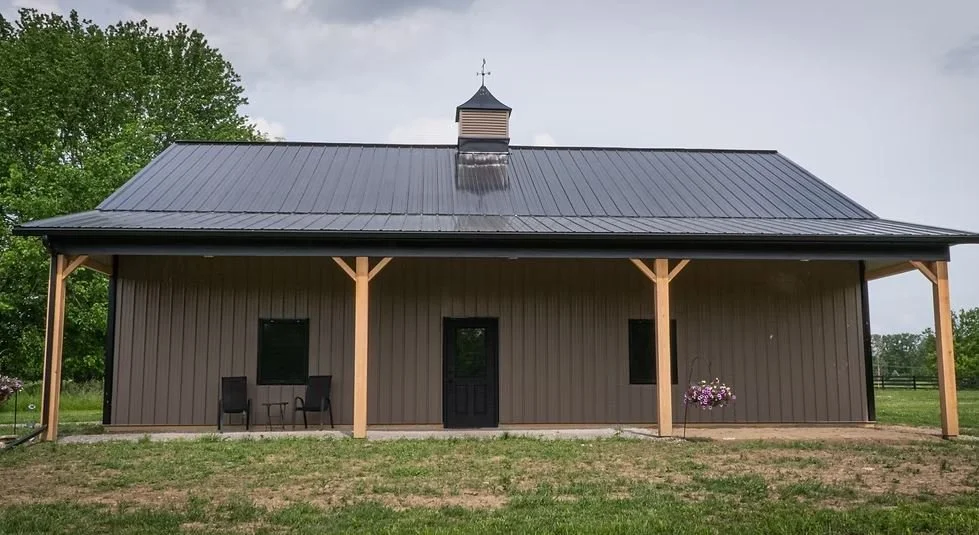 A brown metal building with a metal roof, wooden supports, two small windows, a door, outdoor chairs, and hanging flower baskets, surrounded by green trees and grass.