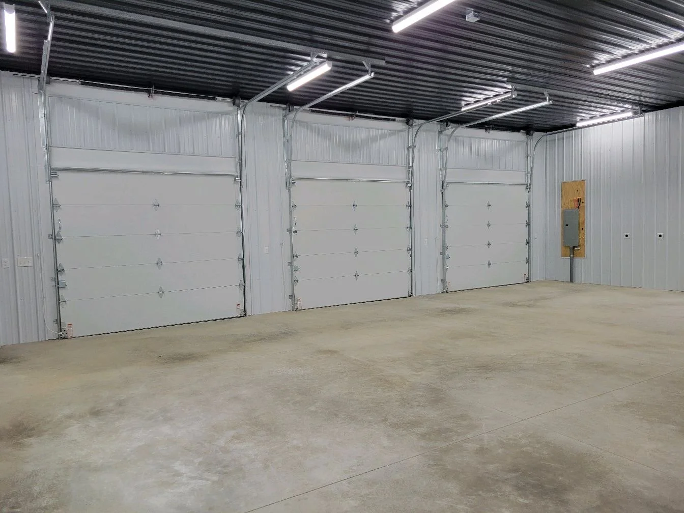 Empty garage with three white sectional roll-up doors, concrete floor, and metal-paneled walls and ceiling.