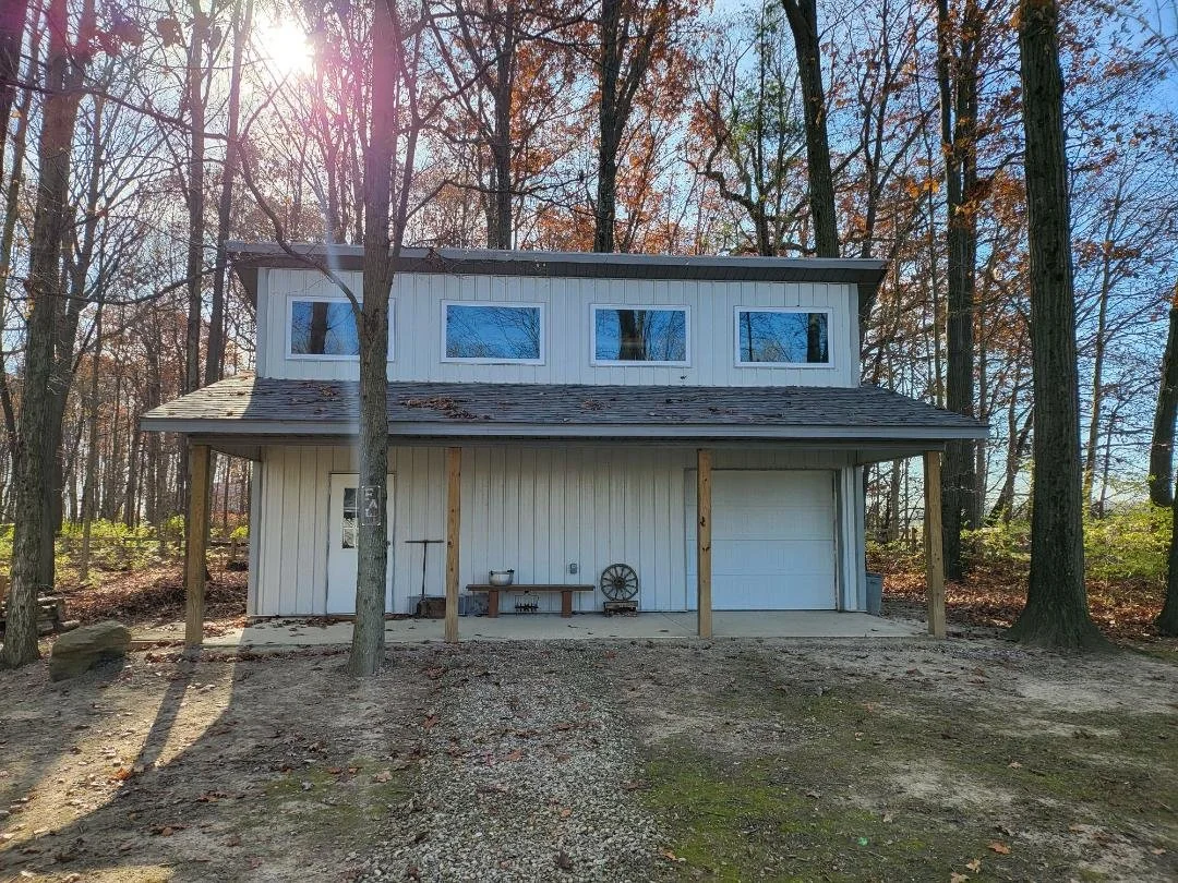A two-story house with white siding and a garage on the right side. The house is surrounded by leafless trees, suggesting fall or winter, with sunlight filtering through the trees.
