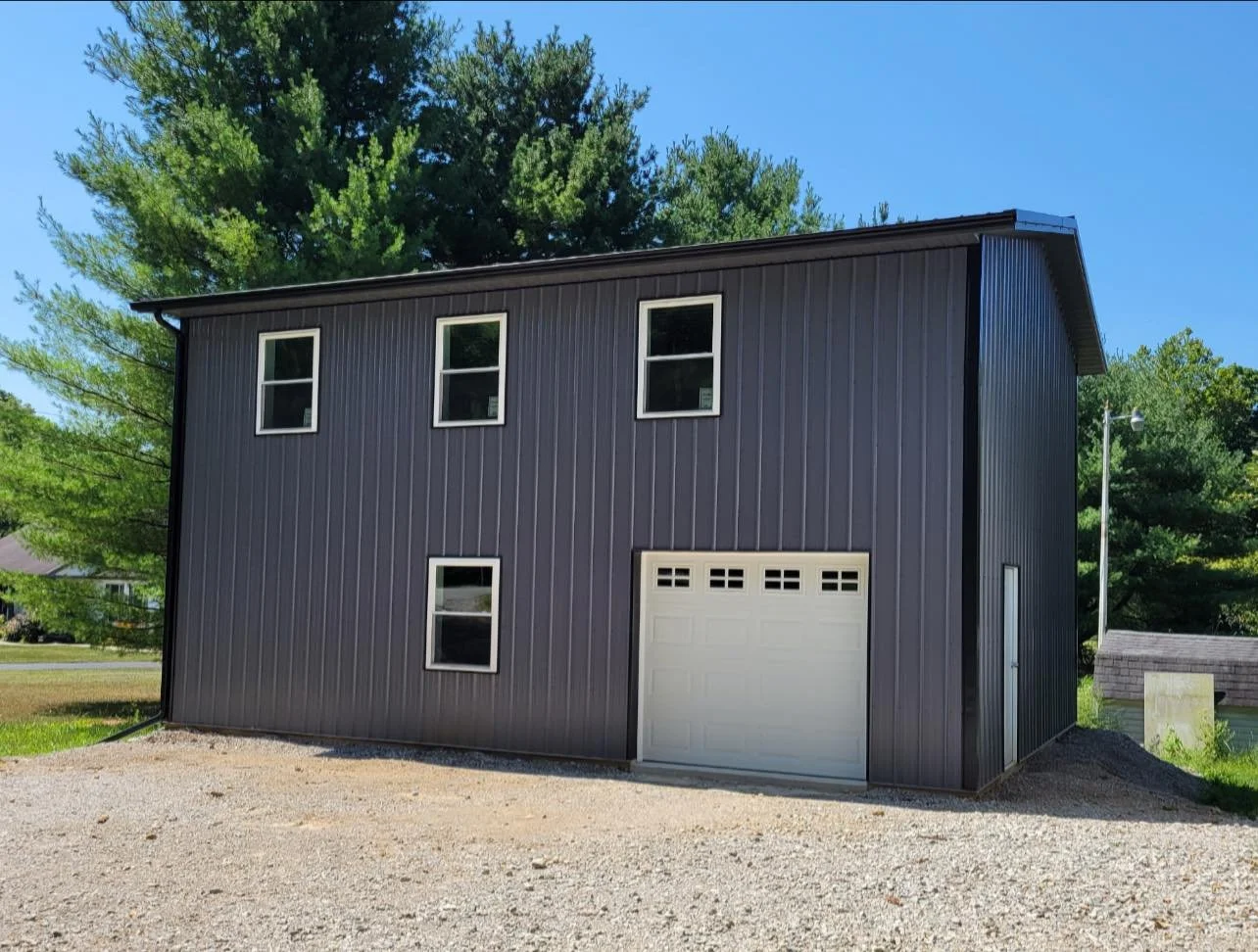 A modern two-story black metal garage with four small windows and a white garage door, set on gravel ground with green trees and blue sky in the background.