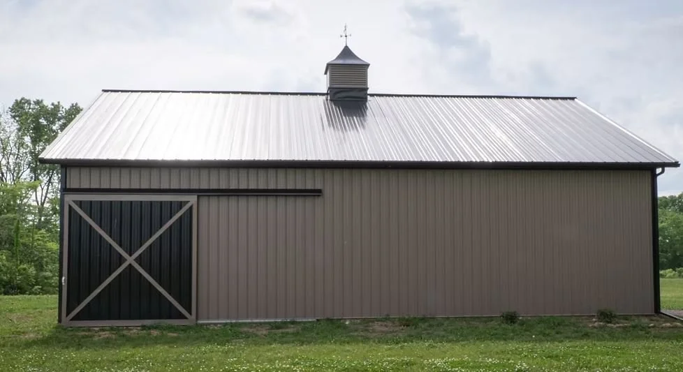 A large gray barn with a metal roof, sliding door with an X-shaped brace, surrounded by green grass and trees, under a partly cloudy sky.