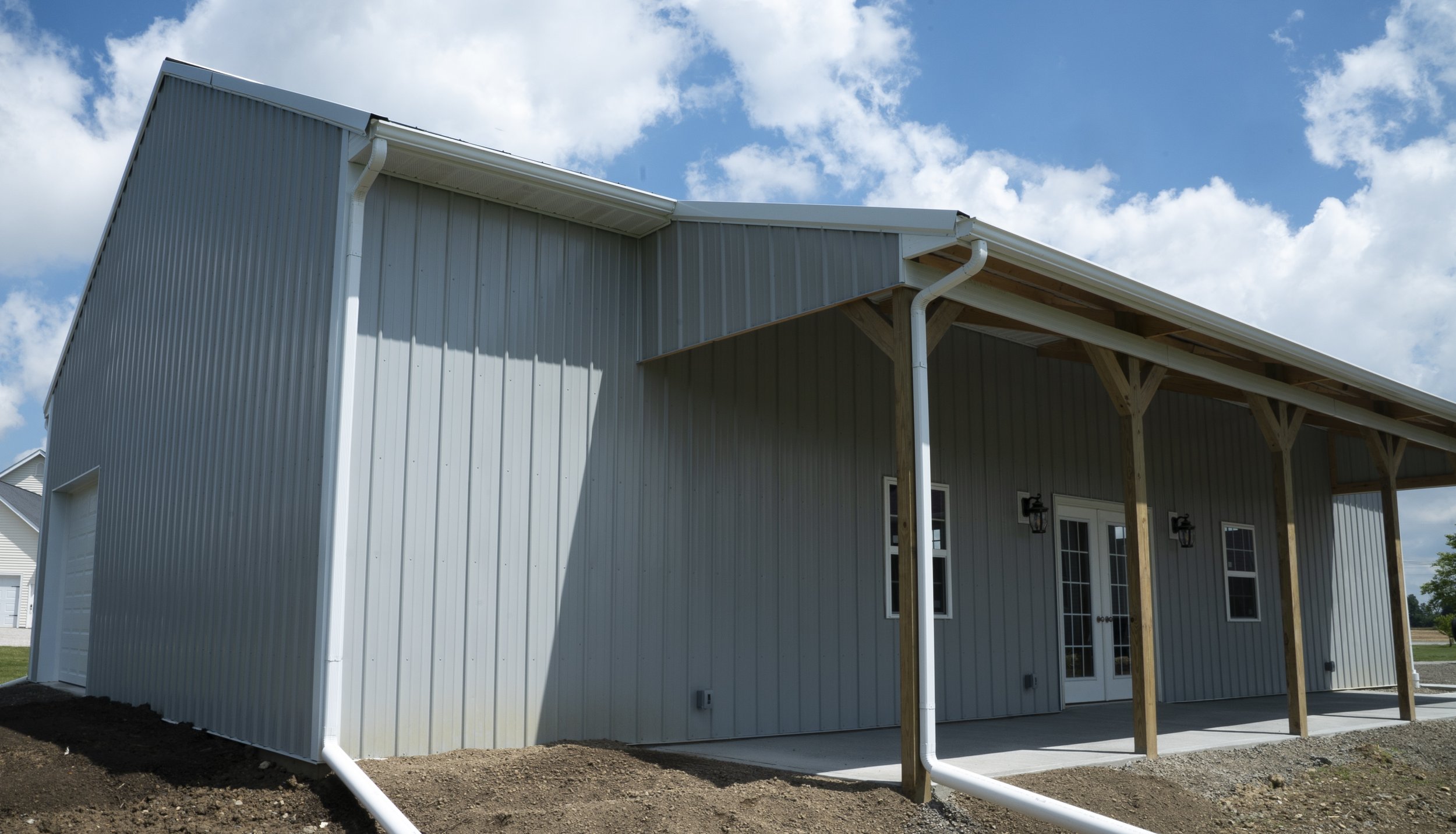 A large metal building with a covered porch area, supported by wooden beams, and a white door with windows, under a partly cloudy sky.
