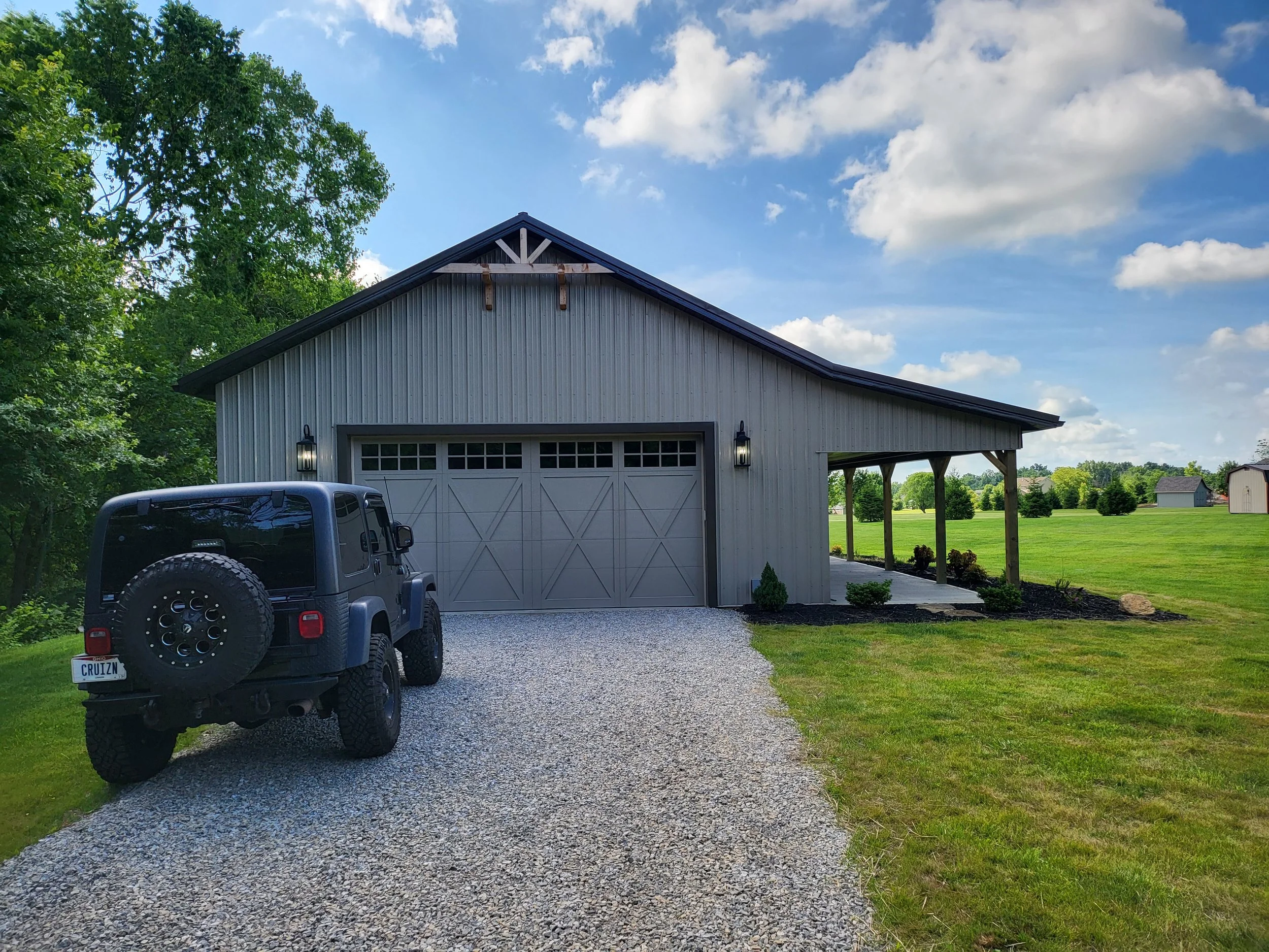 A residential garage with a closed white door, a driveway made of gravel, a black Jeep parked to the left, and a gray barn-style house with a gable roof in the background. The house is surrounded by green grass and trees, and the sky is partly cloudy