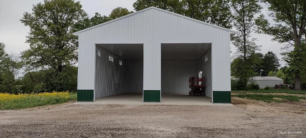 Empty white metal barn with green trim, open entrance, on gravel driveway, with trees and a small building in the background.