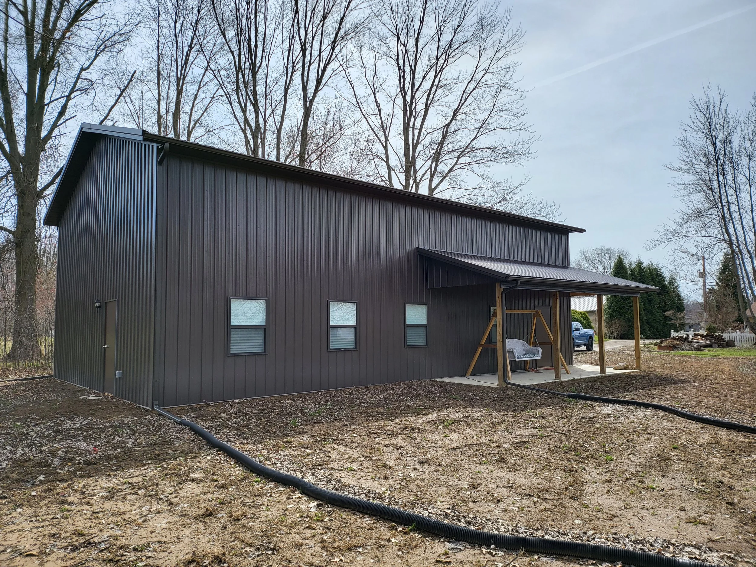A modern black metal building with vertical siding and windows, set in a wooded area with leafless trees and a dirt yard, during daytime.