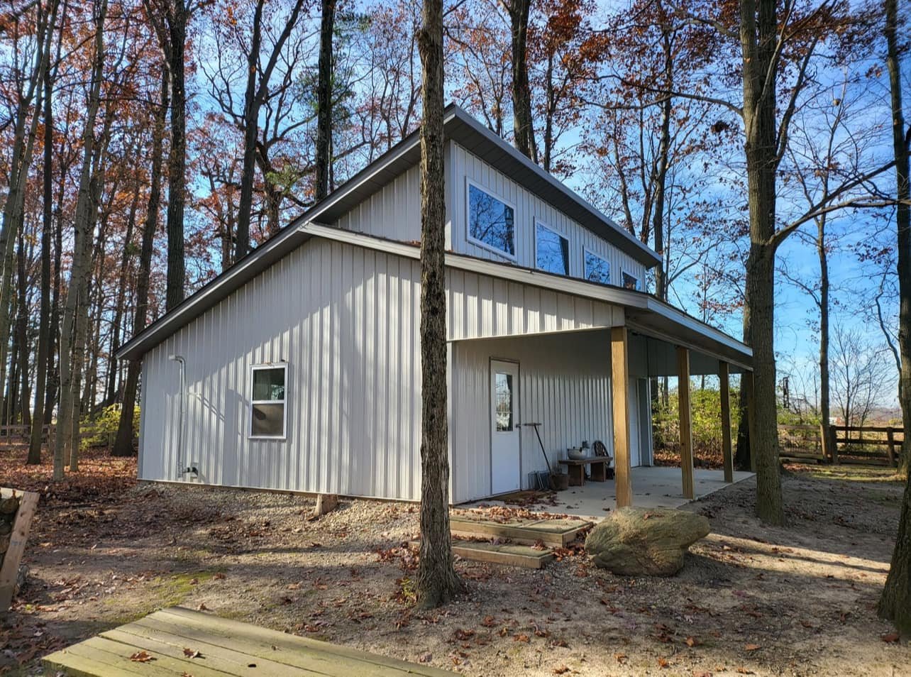 A modern two-story house with white metal siding, surrounded by trees with fall foliage, featuring an overhang porch supported by wooden posts.
