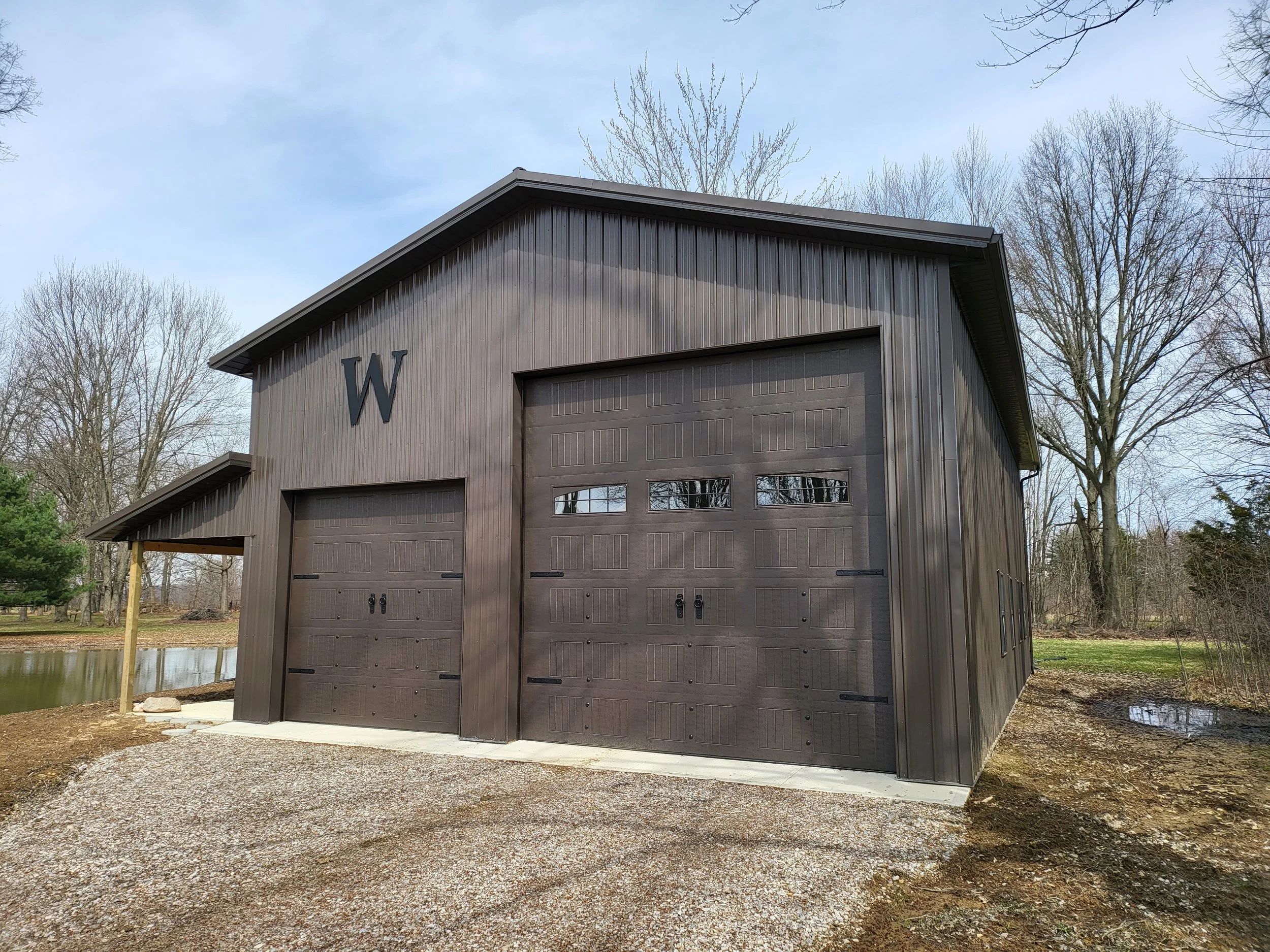 A gray metal building with large garage doors and the letter 'W' on its side, situated outdoors near trees and a small body of water during daytime.
