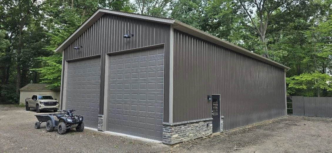A large metal garage with two garage doors, an electric vehicle charging station, a pickup truck, and an ATV parked outside. Surrounded by trees and a gravel driveway.