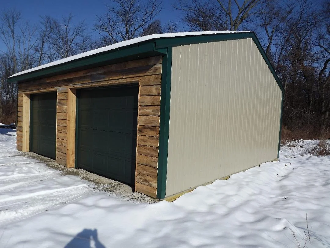 A metal and wood storage shed with two garage doors on snowy ground, trees in the background, and a clear blue sky.