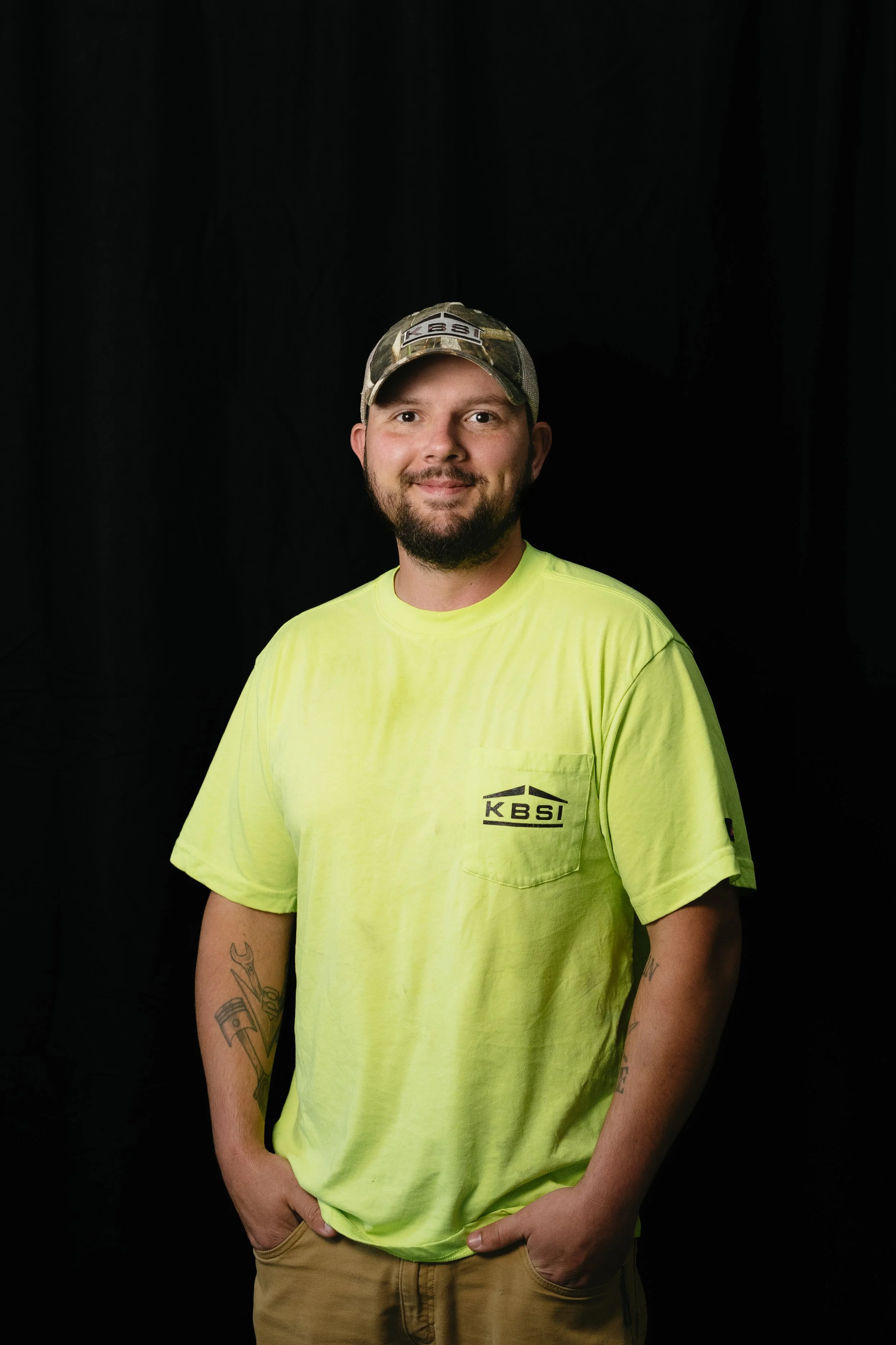 A smiling man wearing a camouflage cap and a neon yellow T-shirt with 'KBSI' printed on it, standing against a black background.