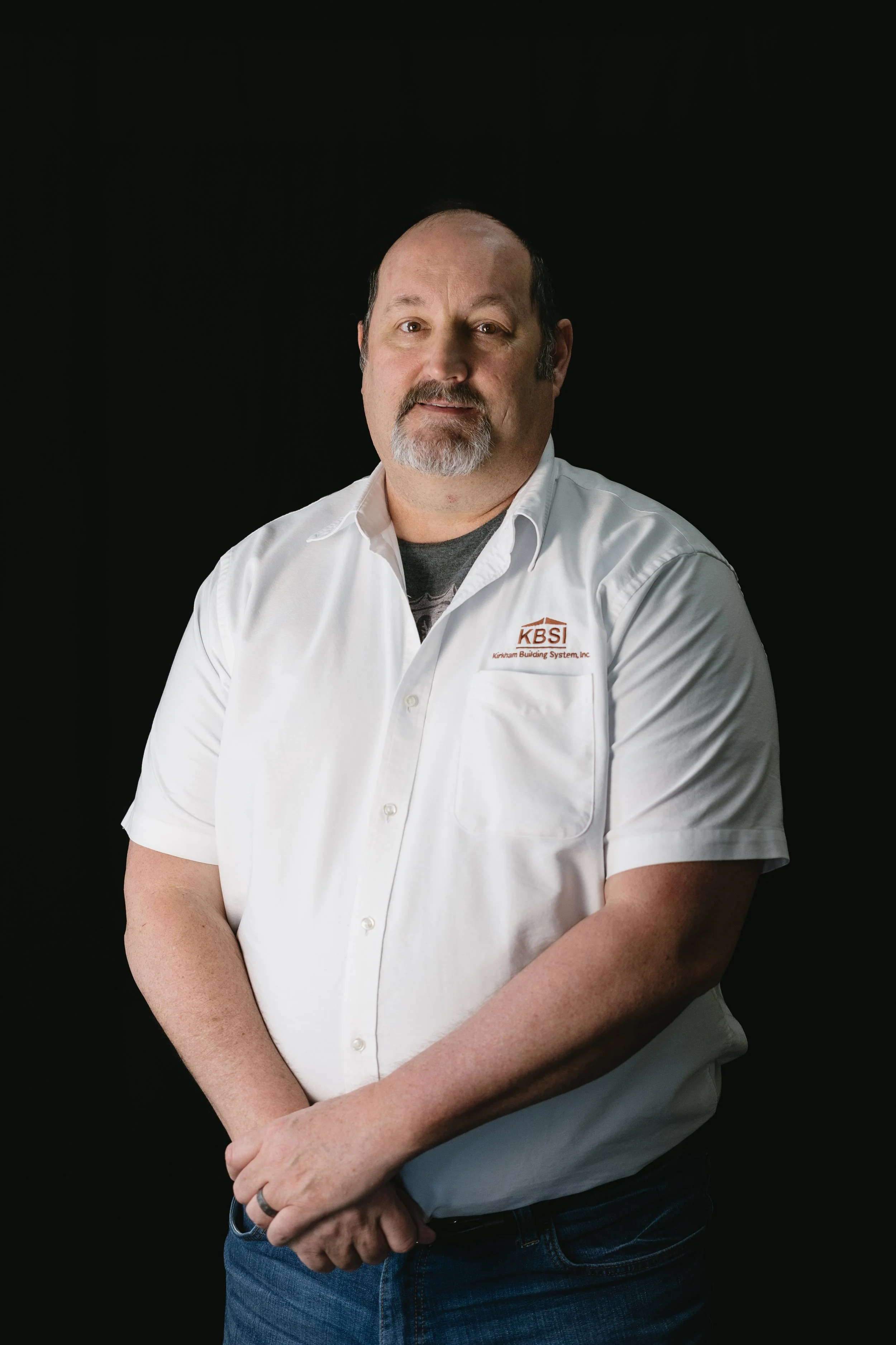 Portrait of a middle-aged man with a beard and mustache, wearing a white shirt with a company logo, standing against a black background.