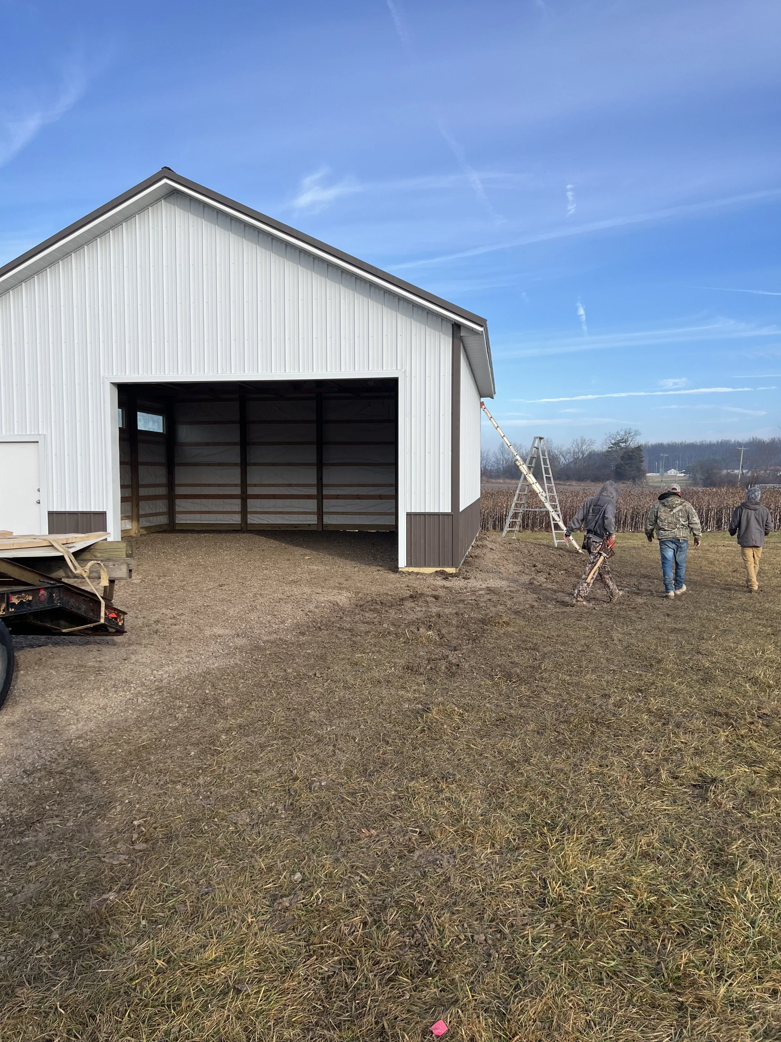A white metal building with a large open garage door, set in a rural area with cloudy blue sky and a few trees in the background. Three people walk away from the camera toward the building, two wearing camouflage clothing, one in a jacket and hat. Th