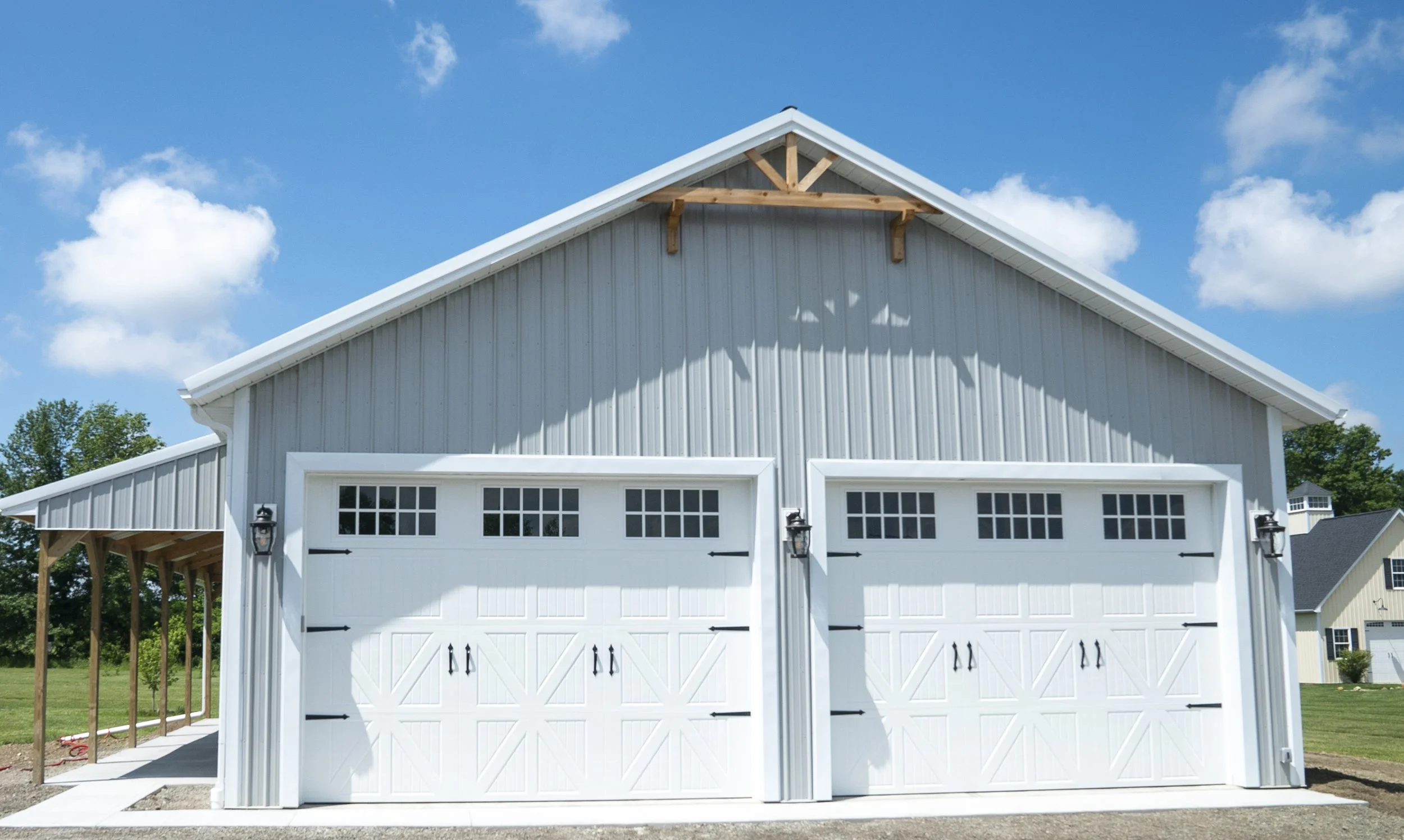 A white garage with double doors, decorative black hardware, and windows, set on a grassy area with a concrete driveway, under a blue sky with some clouds.