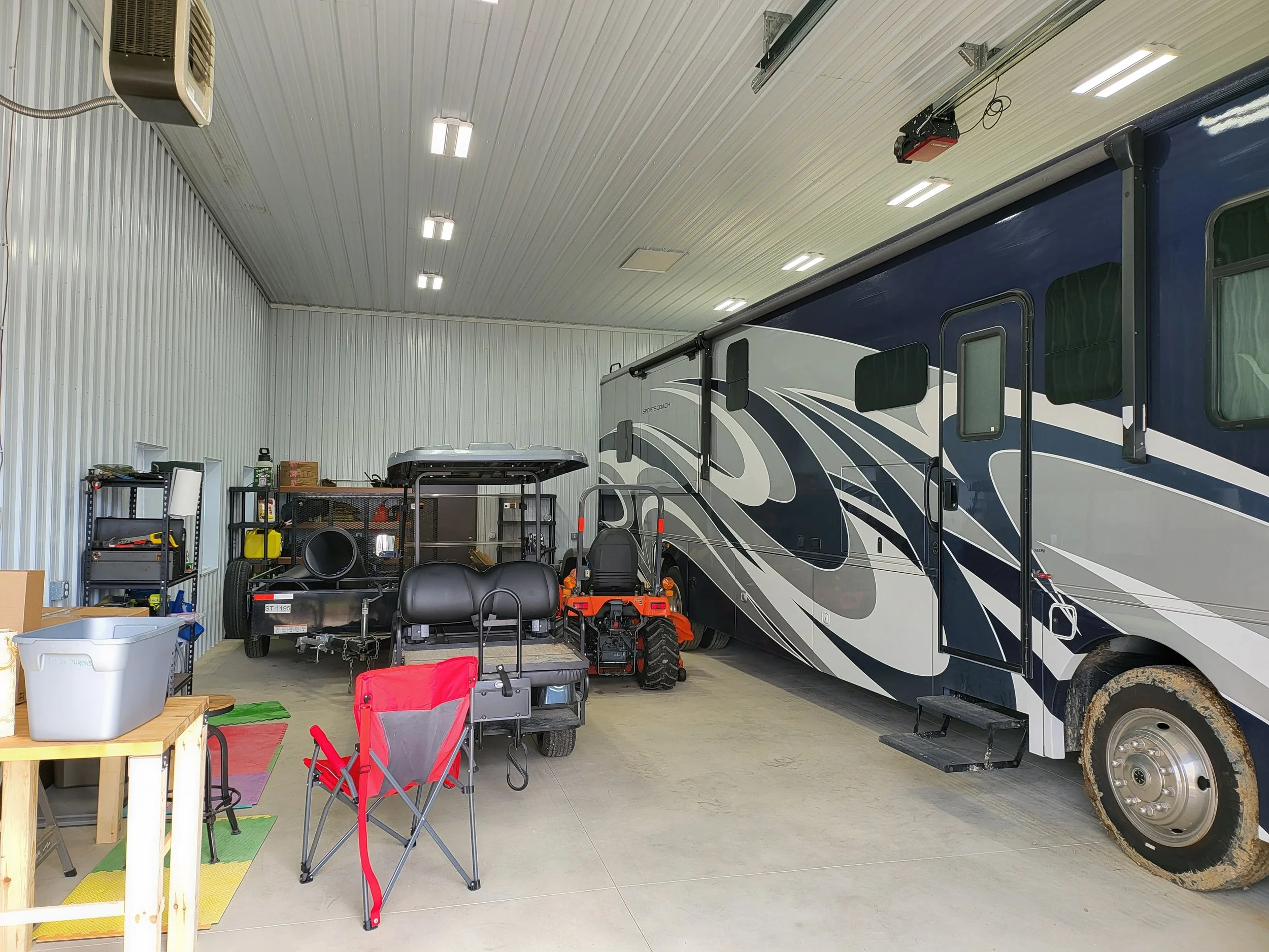An indoor storage garage with a large recreational vehicle (RV) and various outdoor equipment and furniture. The garage has a high ceiling with bright fluorescent lights and metal walls. There are shelves, a small table, and some gardening tools visi